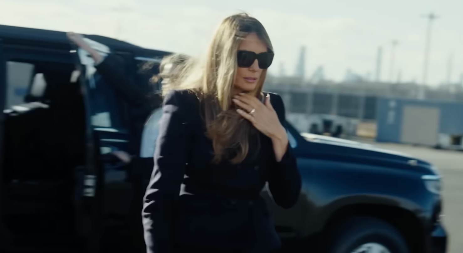 En kvinne i mørk frakk og solbriller går ut av en SUV på et arrangement og justerer håret med én hånd