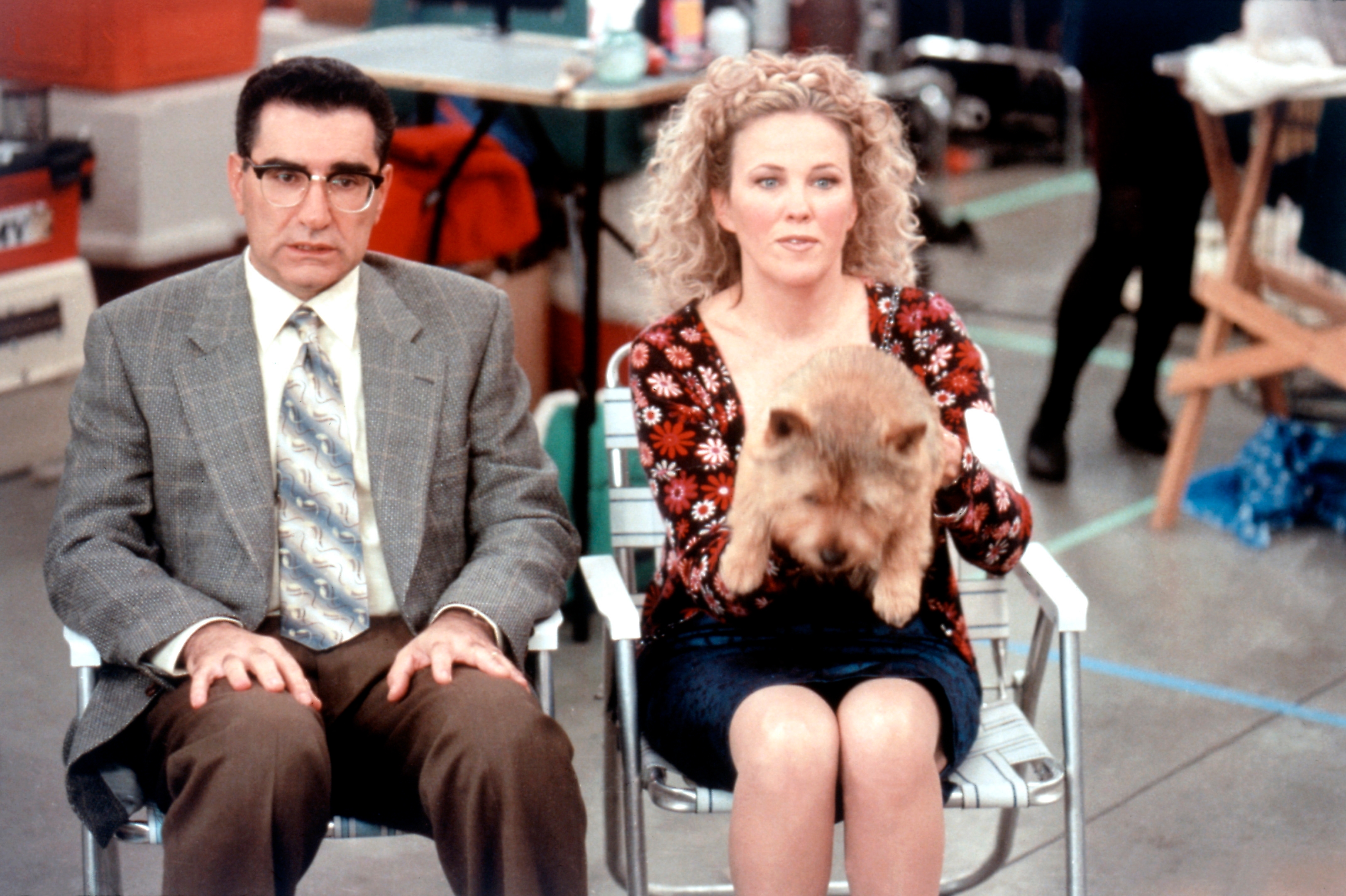 Two people sitting in chairs; the woman holds a small dog. Both appear engaged, possibly on a set