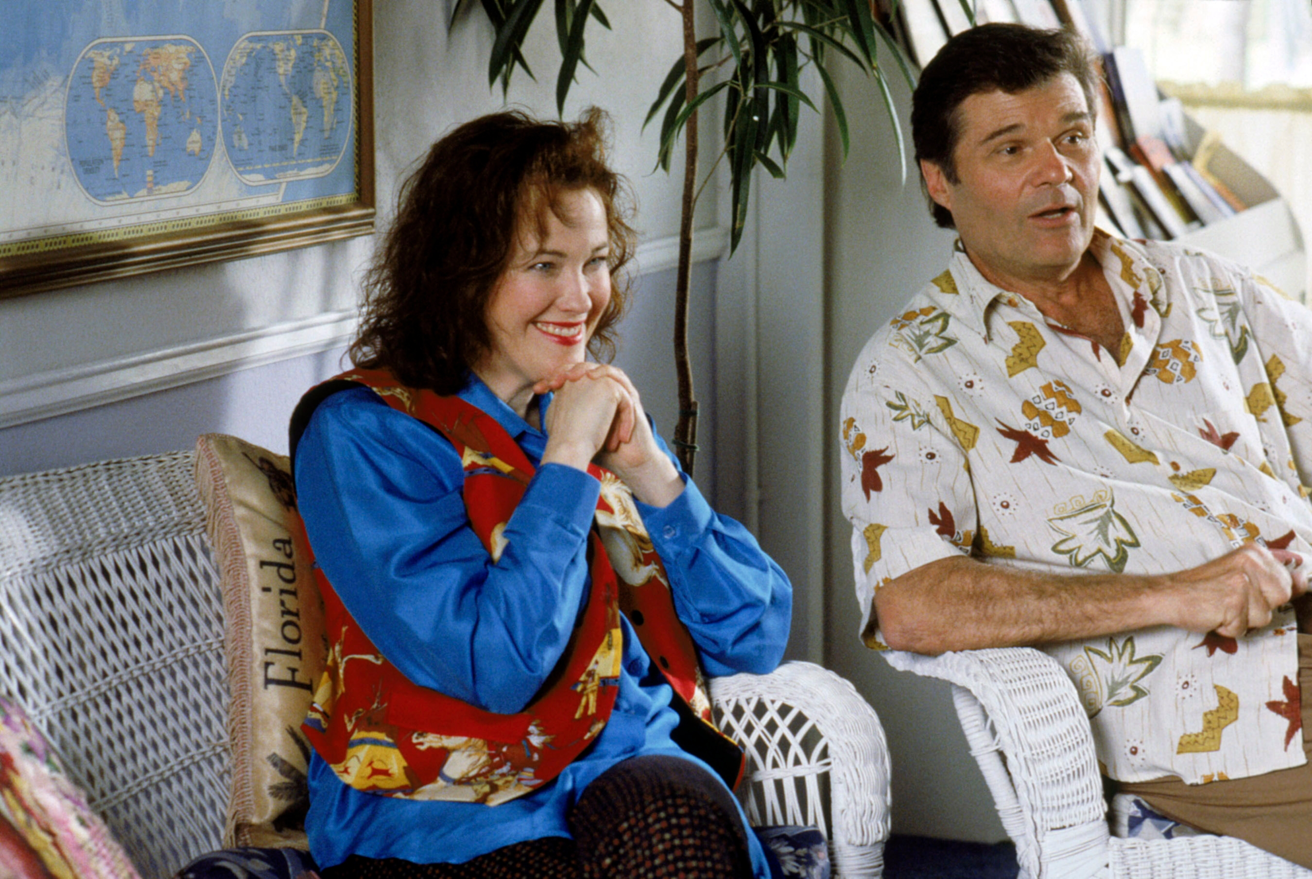 Two people sit on wicker chairs in a casual setting. The woman wears a patterned vest and blue shirt, while the man wears a tropical print shirt