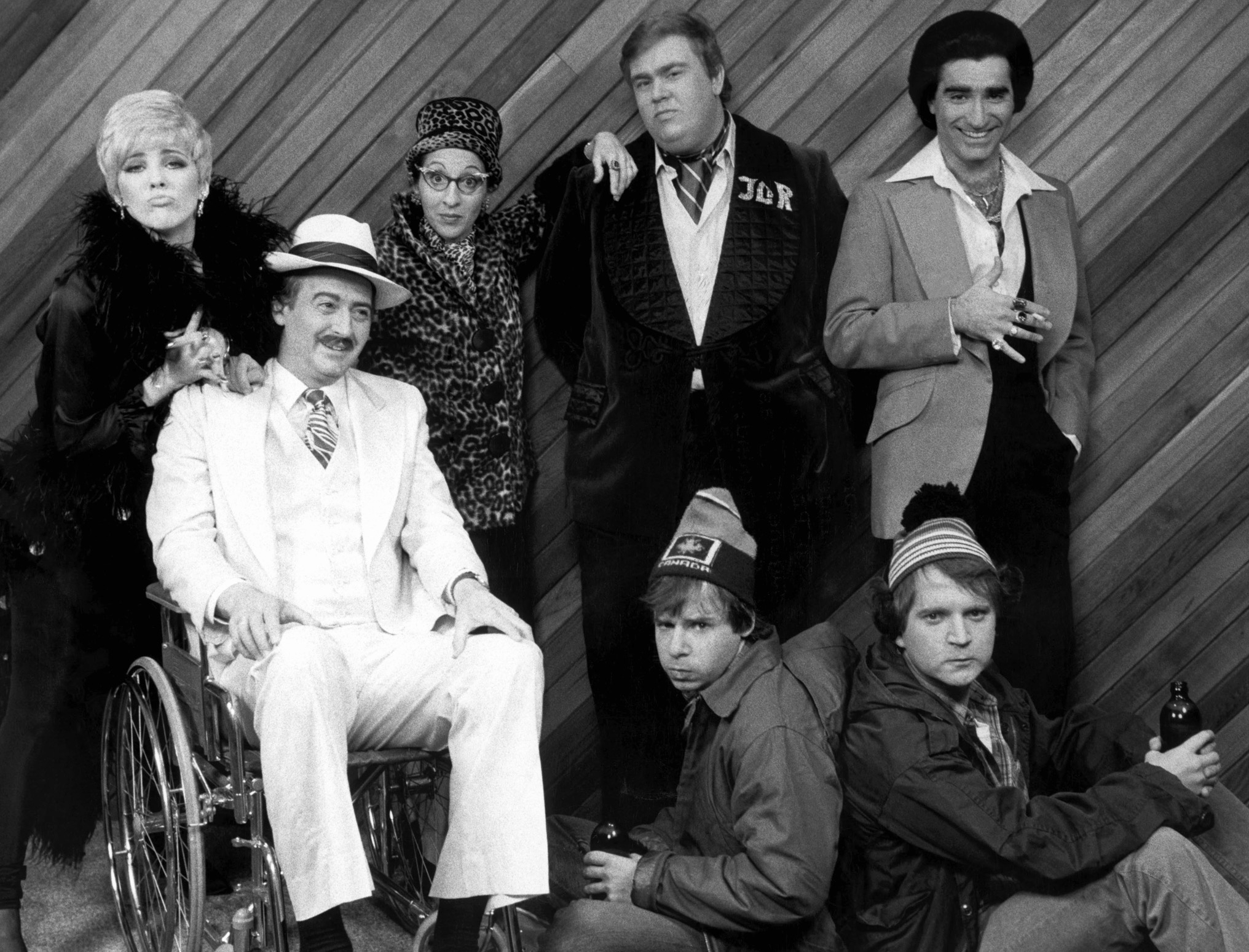 Group of seven adults in comedic poses, dressed in vintage style. One is in a wheelchair wearing a suit. Others wear hats and casual or formal attire