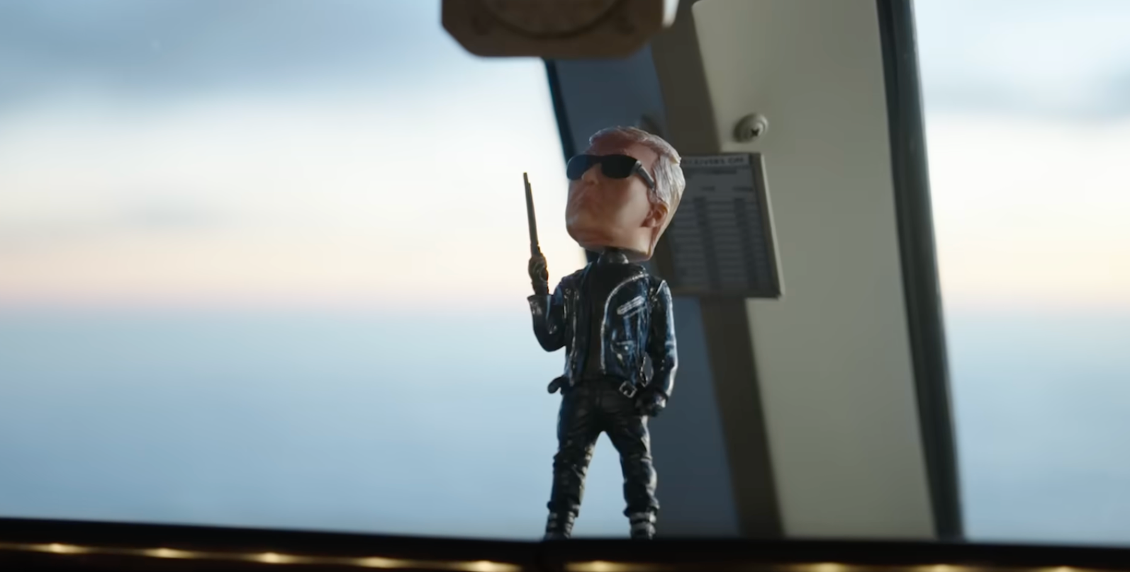 Figur av en person i solbriller og en skinnjakke, som holder en batong, står på et flydashbord mot en vindusutsikt