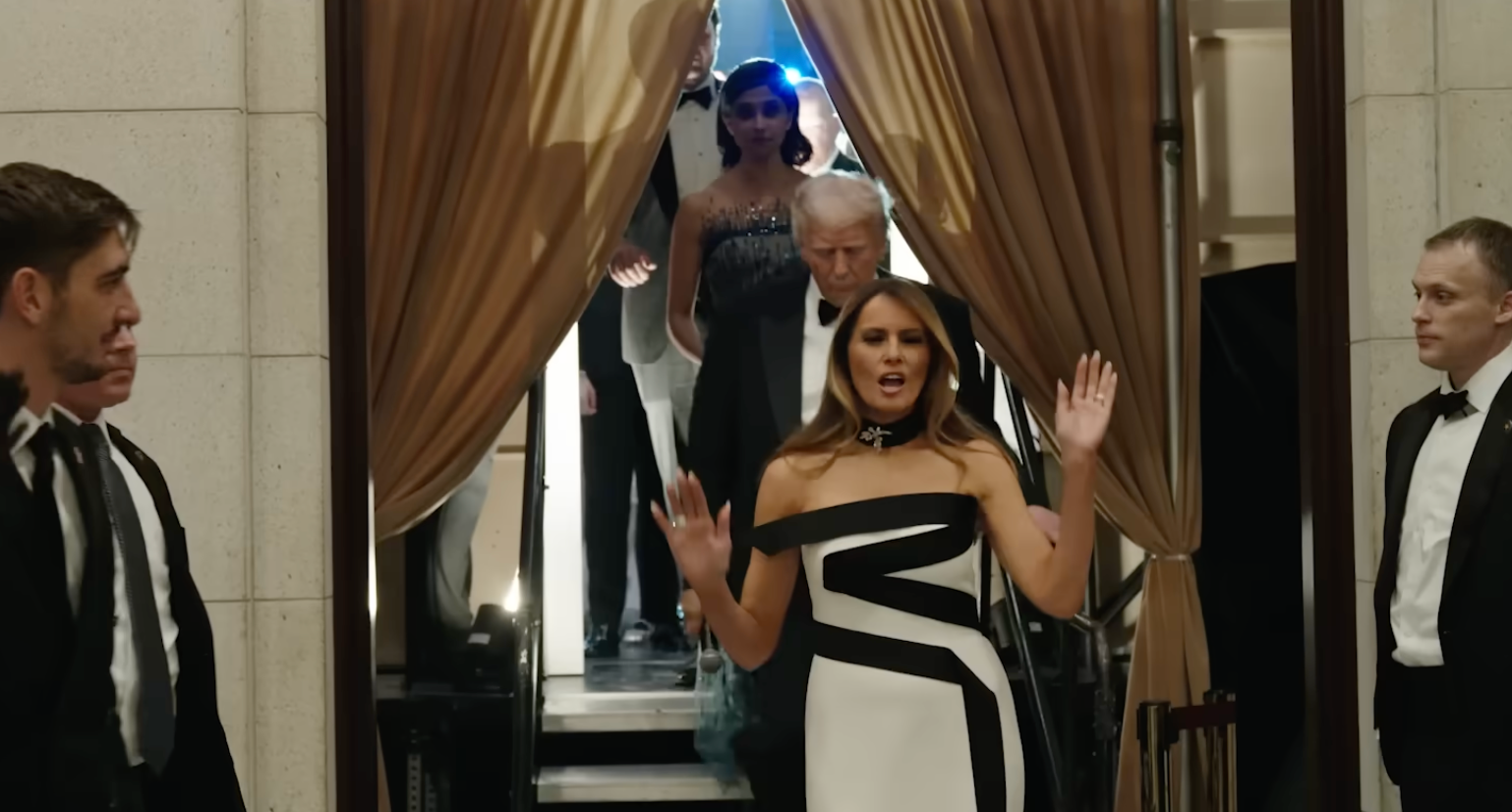 En kvinne i en stilig off-shoulder kjole leder en gruppe ut av en bygning, flankert av menn i formell antrekk