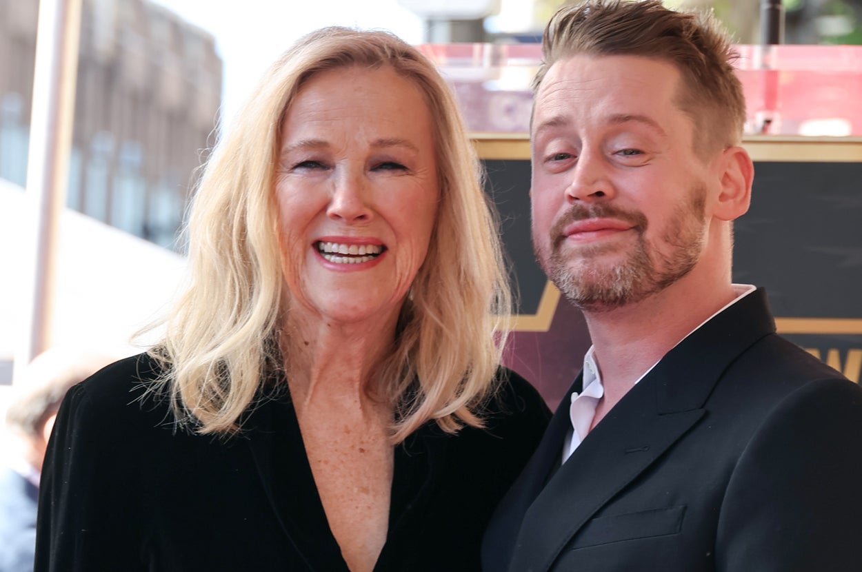 Two celebrities smiling on a red carpet. The woman wears a black outfit while the man is in a dark suit