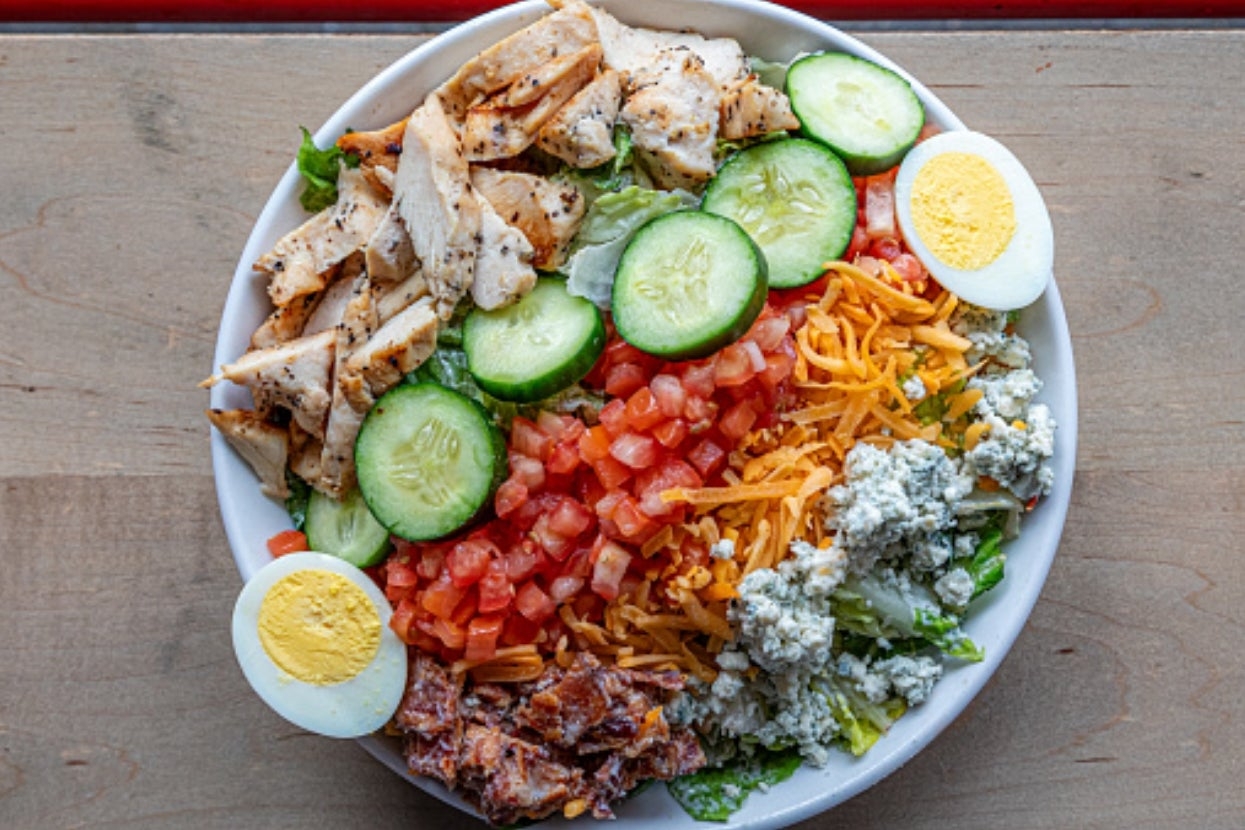 Salad vessel  with grilled chicken, cucumber slices, chopped tomatoes, cheddar cheese, crumbled bluish  cheese, a boiled egg, and bacon bits