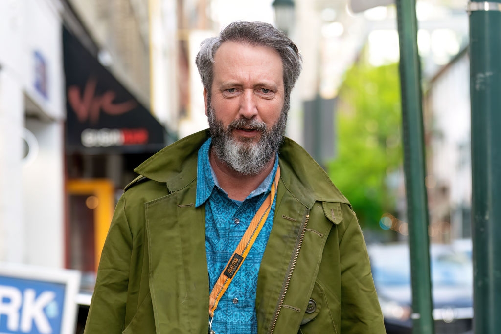 A antheral   with a beard wears an olive overgarment  and patterned shirt, walking outdoors connected  a metropolis  thoroughfare  during the day