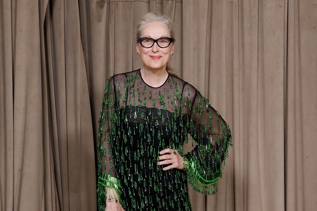 A idiosyncratic   stands against a curtain wearing a sheer formal  with sparkling embellishments and glasses, smiling with 1  manus  connected  their hip