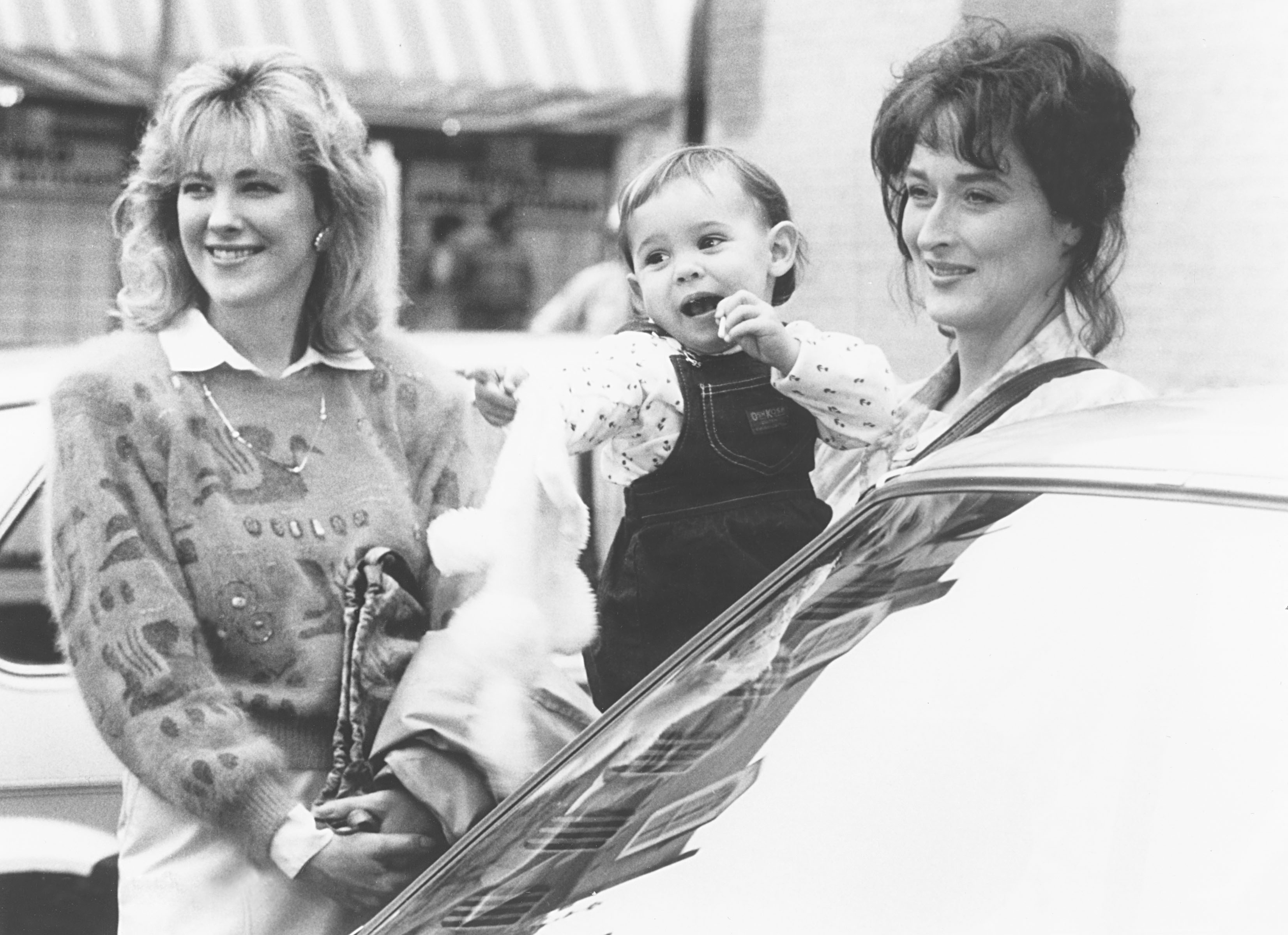 Two women with 1980s hairstyles grin  portion    holding a toddler. The toddler is wearing overalls and holding a stuffed toy