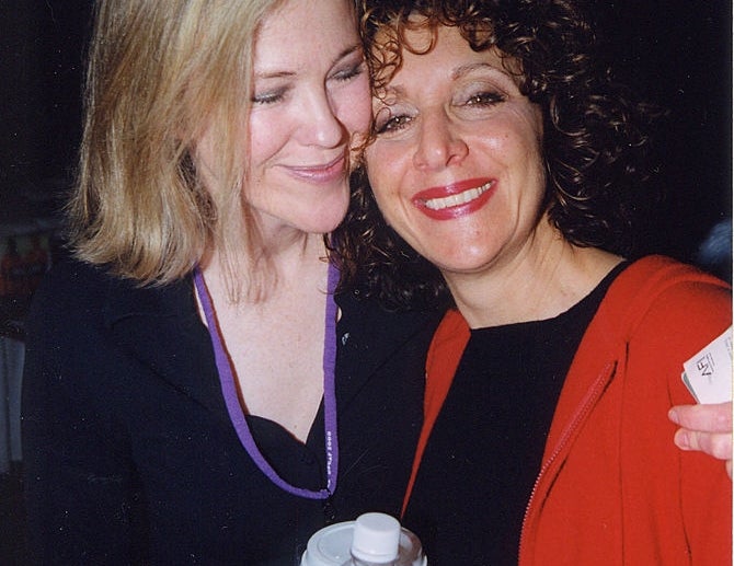 Two radical   smiling intimately  astatine  an event. One has shoulder-length hair, wearing a necklace; the different   is holding java  and wearing a overgarment   implicit    a blouse