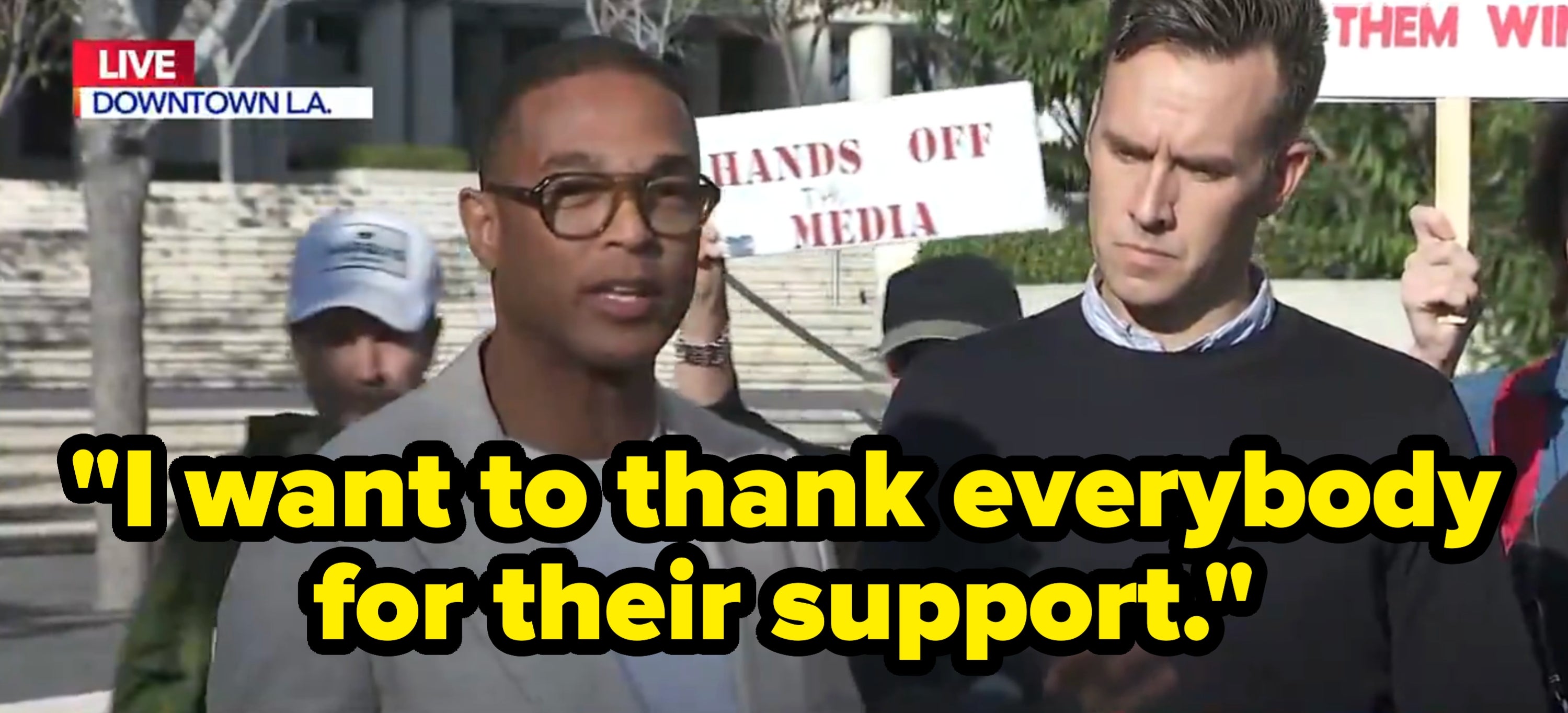 To personer snakker på et pressearrangement med protestskilt i bakgrunnen. Nyhetsbanneret sier: