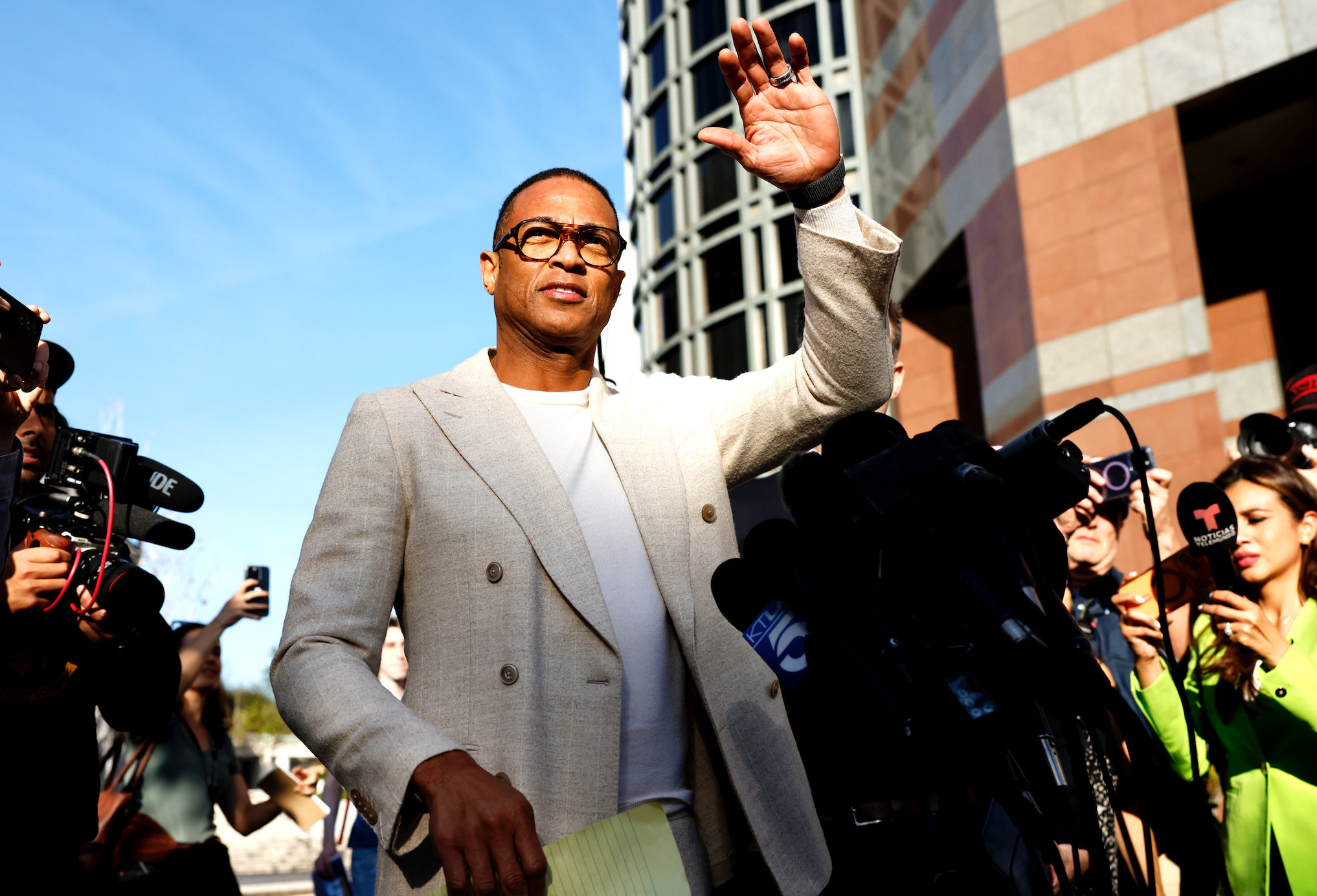 Person i lett dress vinker til fotografer og journalister utenfor en bygning, med kameraer og mikrofoner som fanger scenen