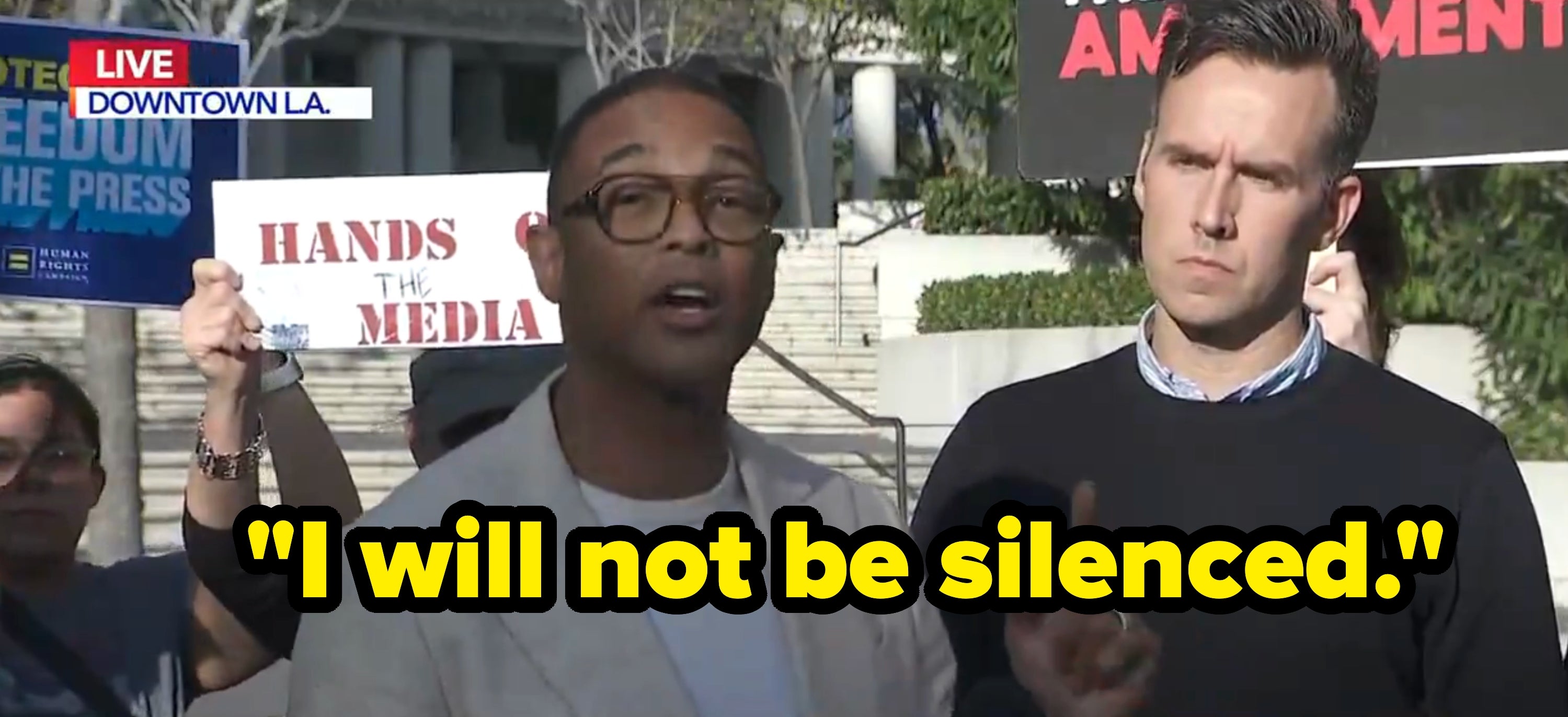 En mann i blazer henvender seg til media på en pressebegivenhet, med en annen mann ved siden av ham. Et skilt med teksten «Hands Off the Media»; er synlig bak dem