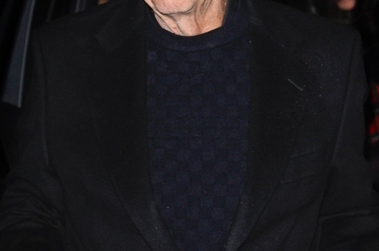 A person wearing glasses and a dark suit exits a vehicle, holding a pen with someone in the background