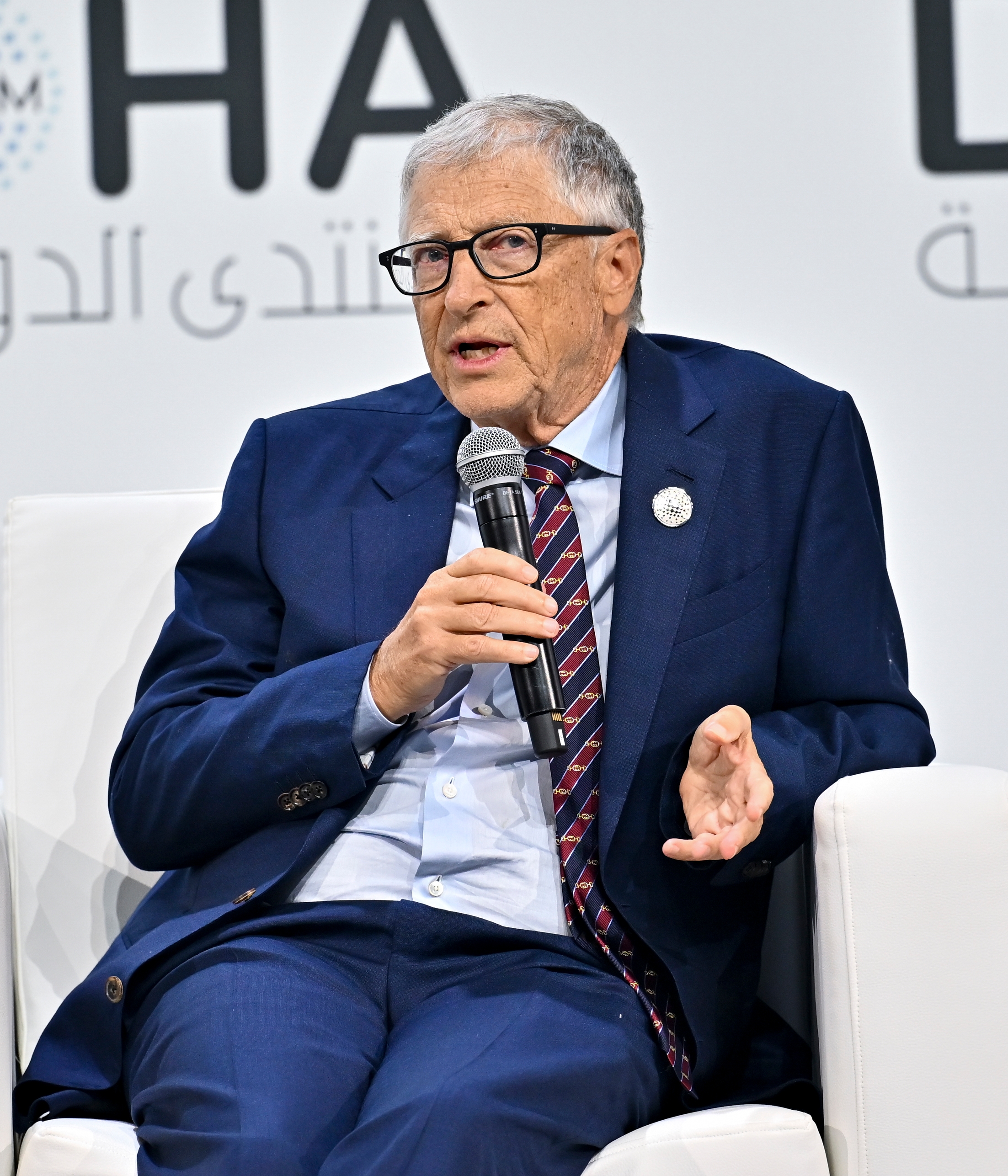 Bill Gates i dress, sittende og holder en mikrofon, snakker ved en formell begivenhet mot et bakteppe med delvis tekst