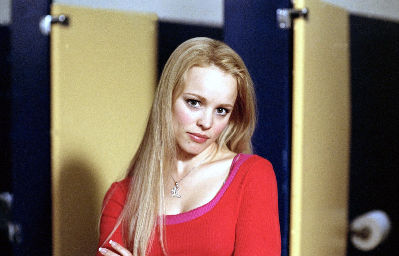 Rachel McAdams with long hair in a red top stands in front of open bathroom stalls, looking directly at the camera