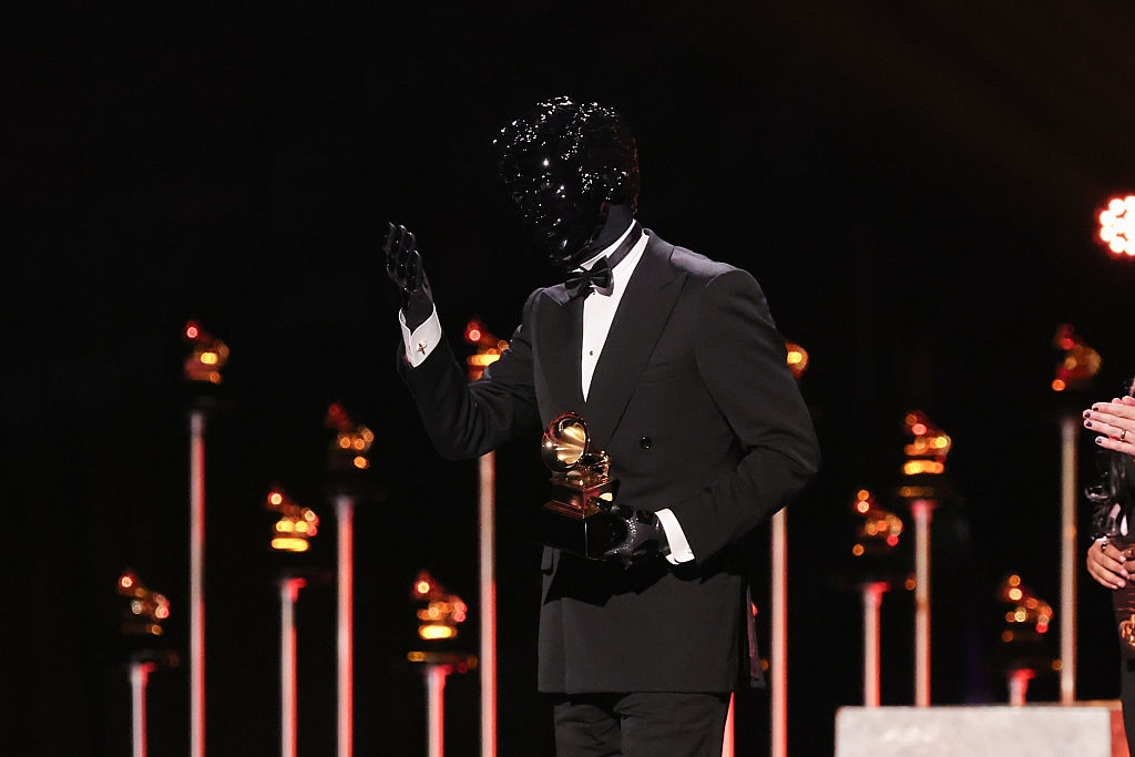 A idiosyncratic   successful  an inventive, glossy achromatic  disguise  and suit   holds a Grammy grant  connected  stage, surrounded by Grammy statuettes