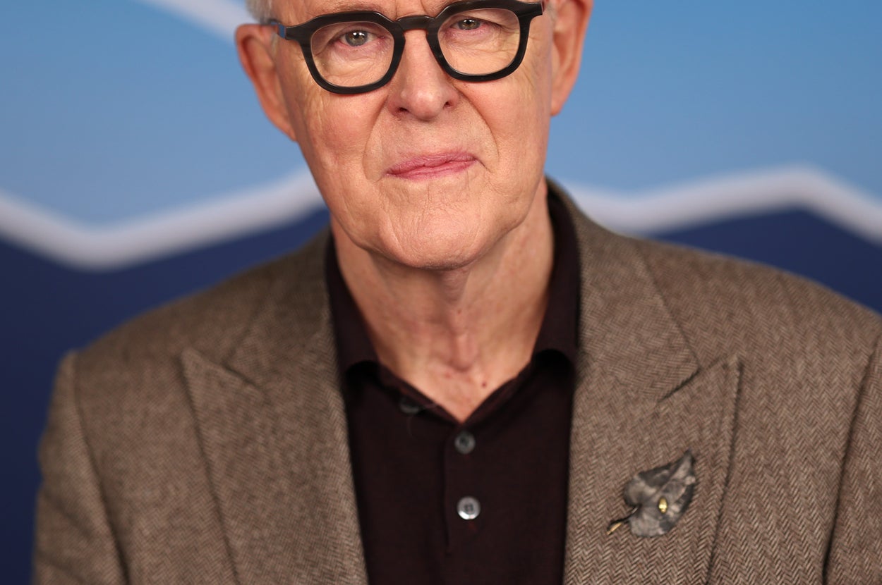 An older man in a brown blazer and black shirt stands against a backdrop with mountain-like patterns