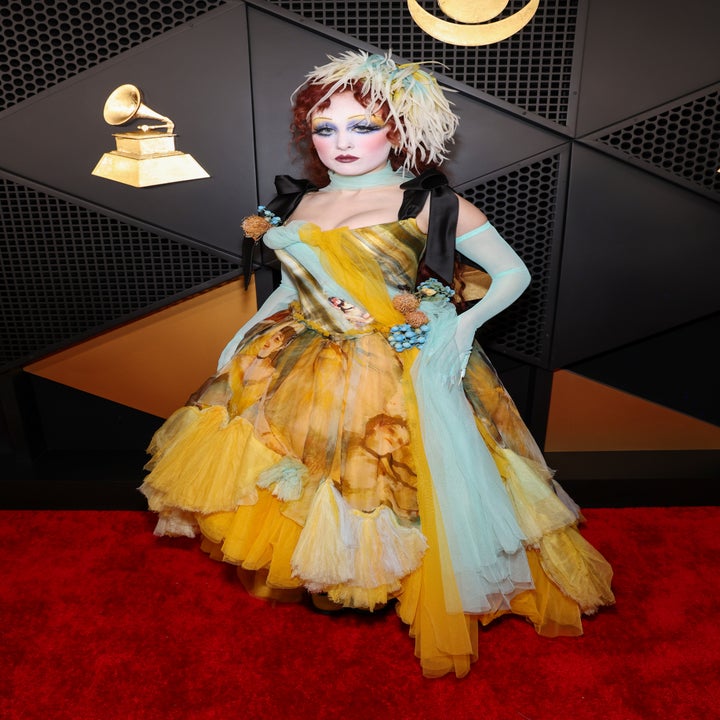 Person on red carpet in a floral patterned gown with glove and feathered headpiece, posing in front of a Grammy backdrop