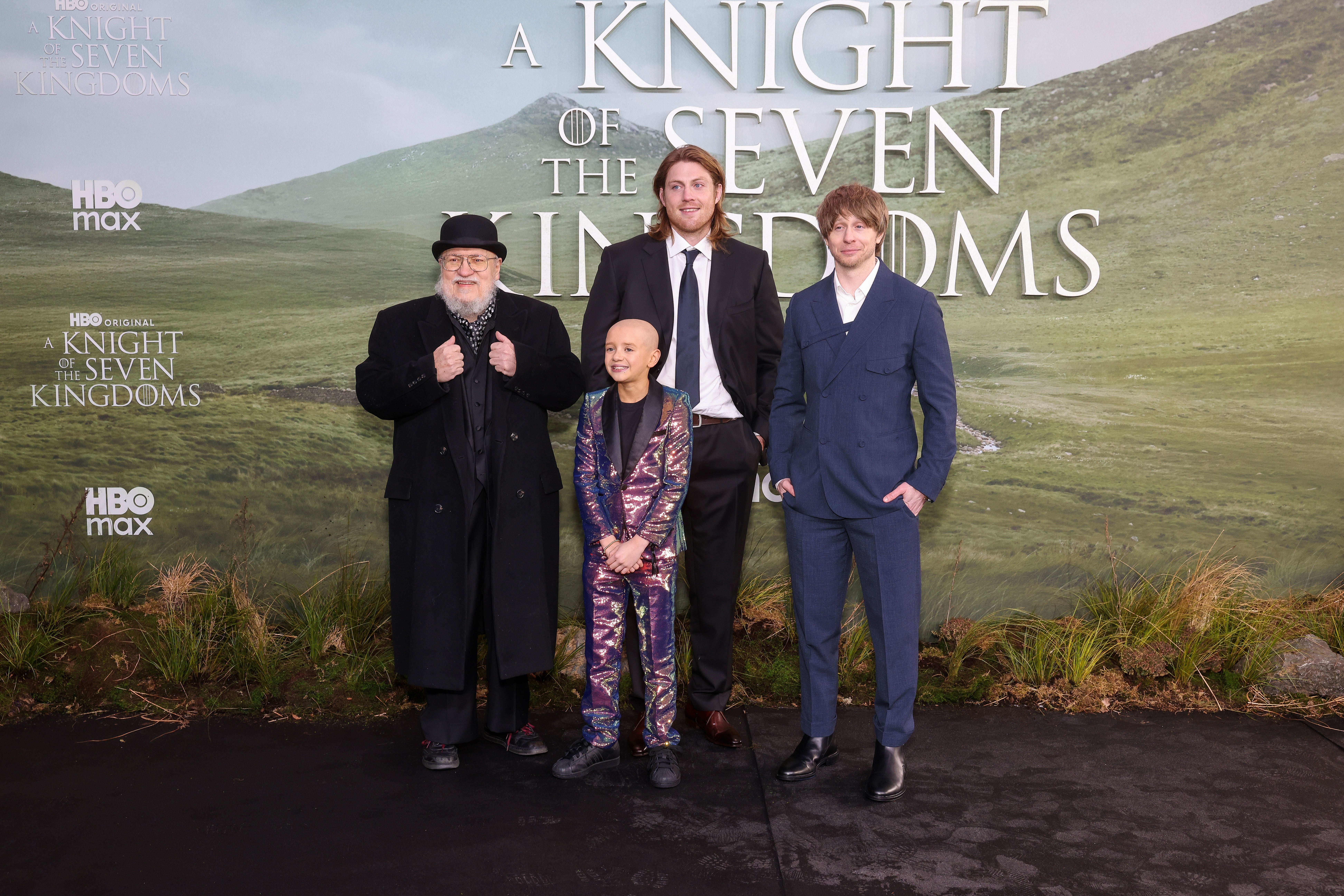 Four people on a red carpet for an HBO Max event, with one in a patterned suit and the others in formal attire, in front of a backdrop displaying the title "A Knight of the Seven Kingdoms."