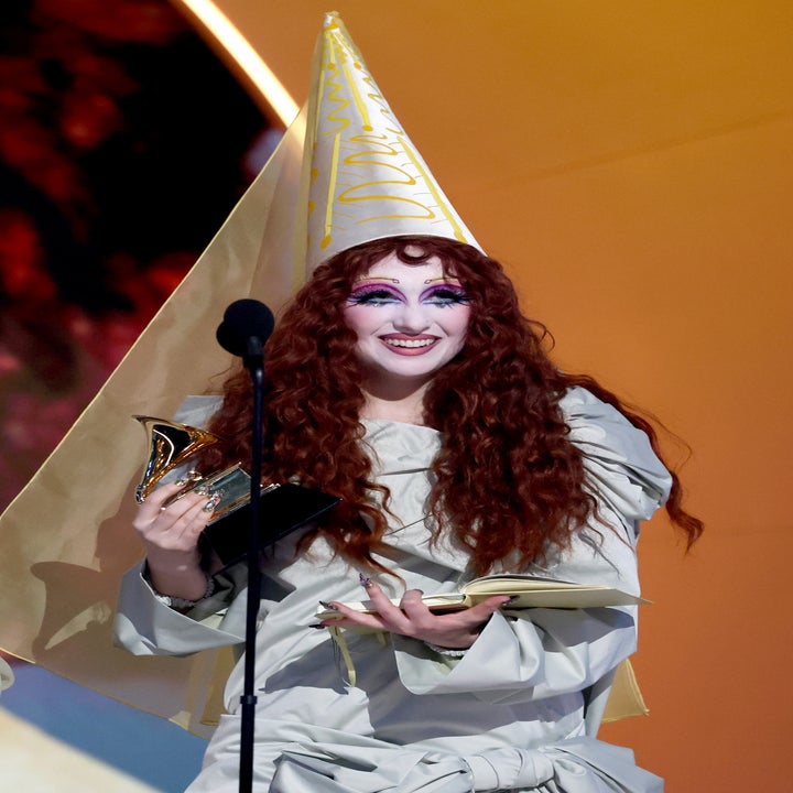 Person with long, wavy hair in a pointed hat and flowing outfit speaks into a microphone, holding an award and notebook on stage