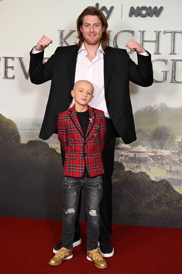 Two individuals pose on a red carpet. The adult, in a suit, flexes. The child wears a plaid blazer and sneakers