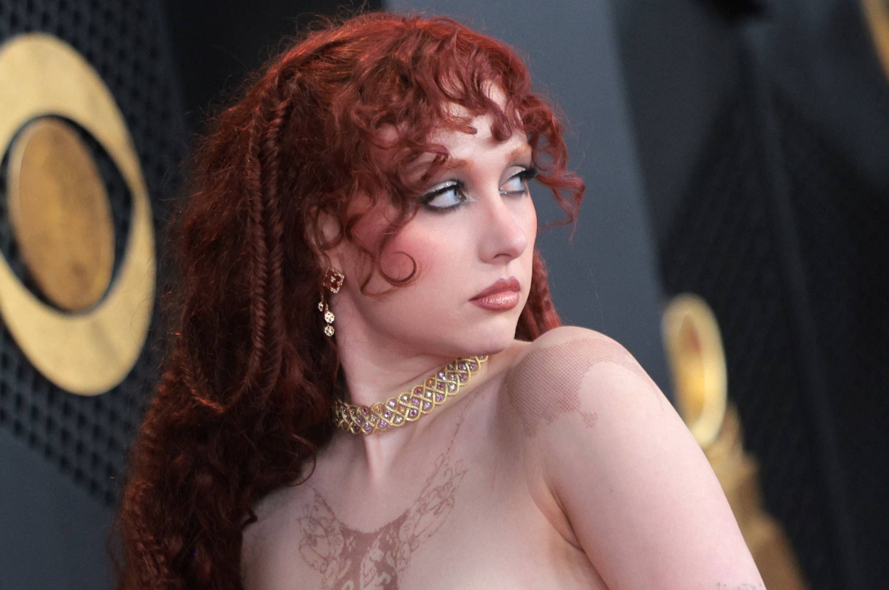 Person with intricate hairstyle in formal attire looks to the side at a glamorous event, showcasing elegant jewelry and detailed outfit pattern