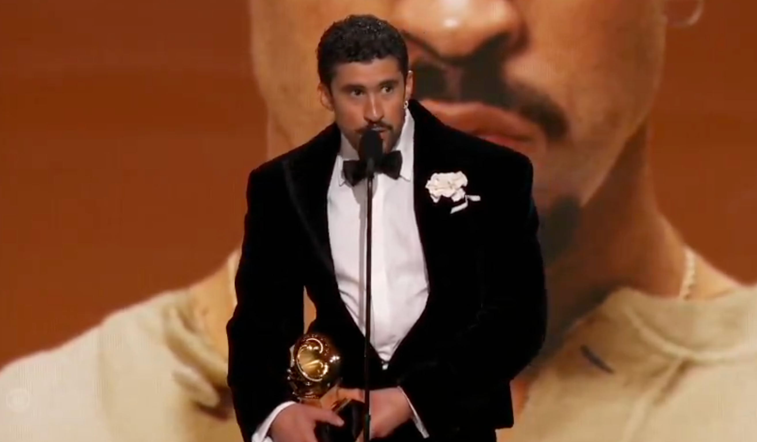 Person successful  ceremonial  attire holding an grant  statue, speaking astatine  an lawsuit   with a ample  representation  successful  the background