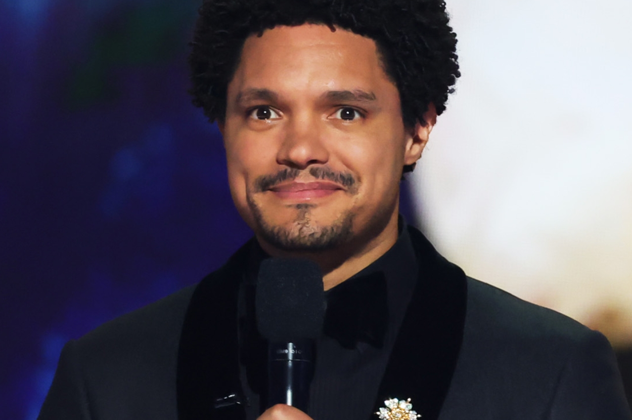 Person holding a microphone, wearing a dark suit with a floral pin, in front of a blurred background