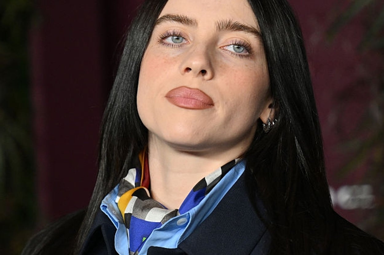 Person in formal attire with sleek long hair, wearing a patterned scarf over a coat, posing at a celebrity event