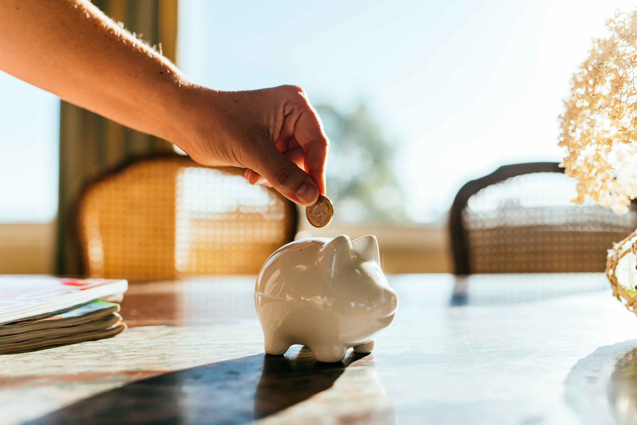 Hand placing coin into a ceramic piggy bank on a sunlit table, conveying a sense of saving or financial planning