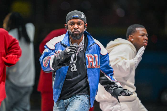 A performer connected  signifier    wearing a leather overgarment   and jeans, holding a microphone with dancers successful  the background