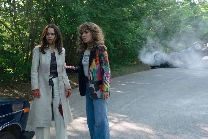 Two people stand on a road, appearing concerned. A car is flipped over and smoking in the background