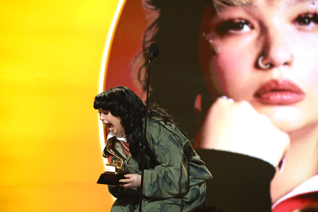 Person connected  signifier    successful  oversized covering  holds a Grammy, expressing joy, with a ample  representation  of themselves successful  the background