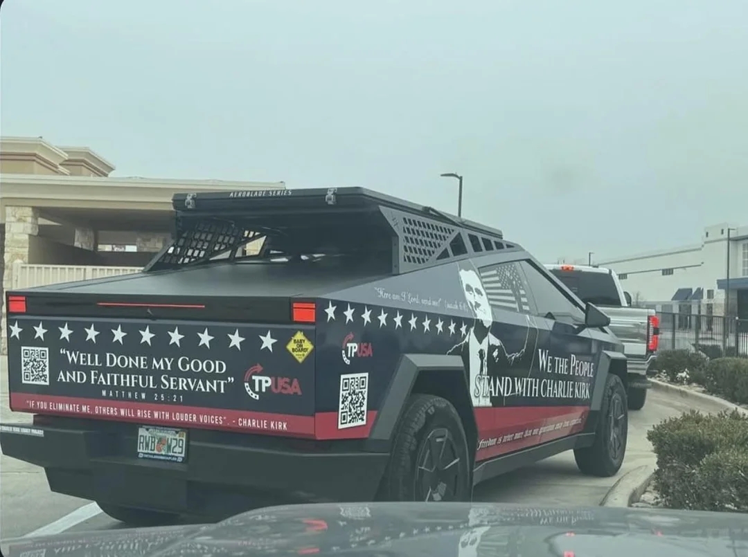 Modified motortruck  with matte decorativeness  displays slogans and logos, including "We the People," "Charlie Kirk," and biblical reference, seen from the rear connected  the road