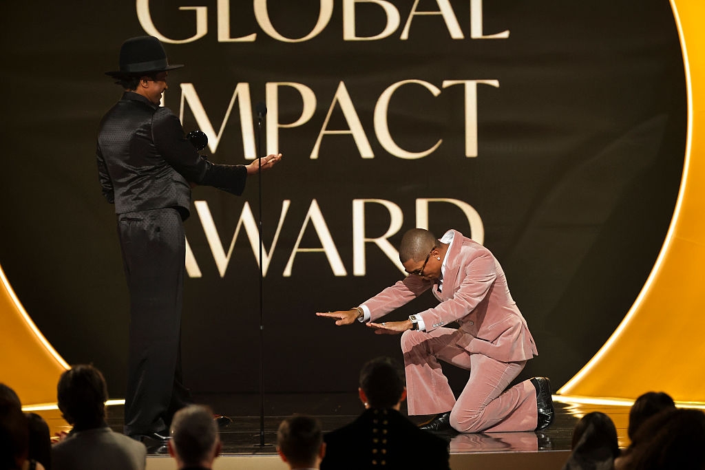Two men successful  ceremonial  attire connected  a stage, 1  kneeling with arms outstretched and the different   lasting  with arms open, astatine  a Global Impact Award event
