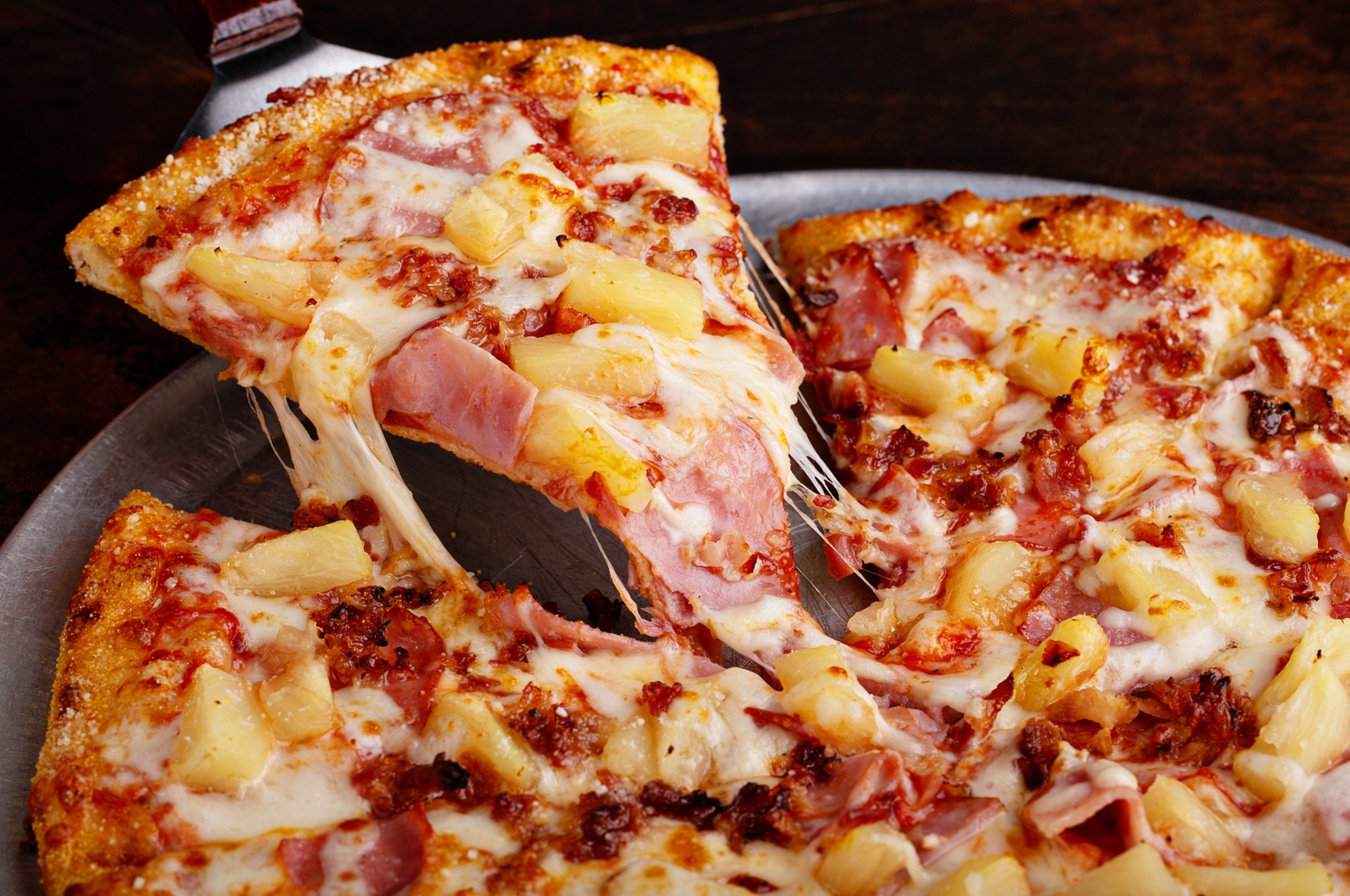 A close-up of a Hawaiian pizza with melting cheese, pineapple chunks, and ham slices on a metal tray
