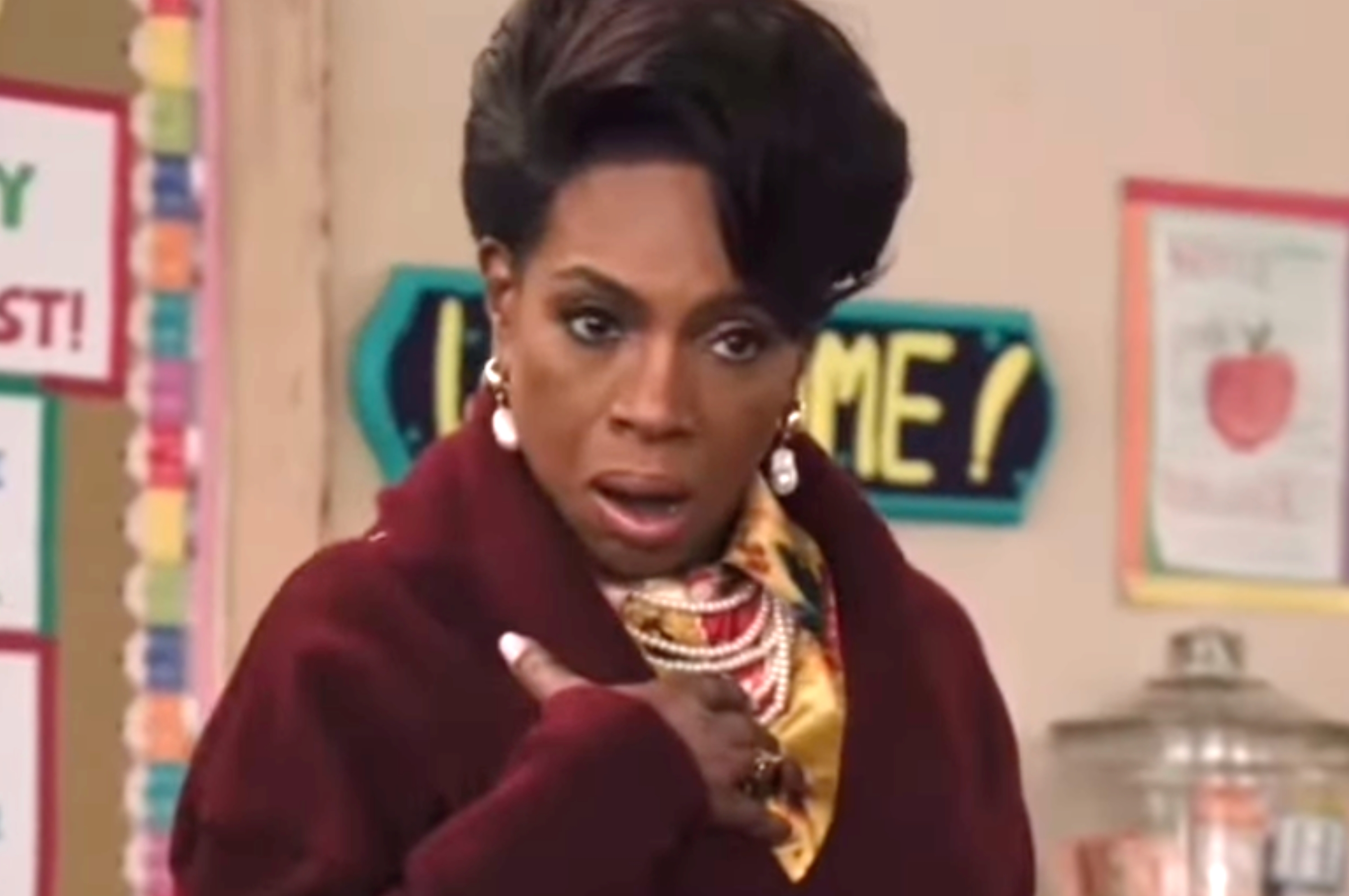 Person in elegant attire with layered necklaces, earring, and styled hair, appearing surprised in an indoor setting with educational posters