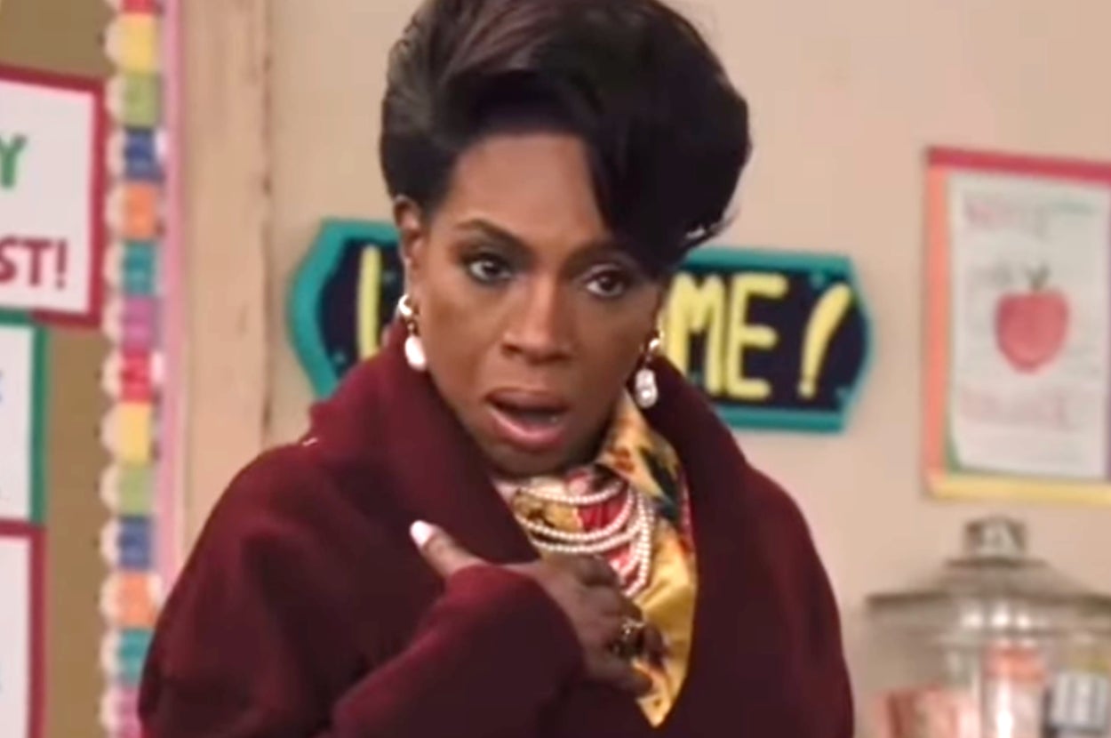 Person in elegant attire with layered necklaces, earring, and styled hair, appearing surprised in an indoor setting with educational posters