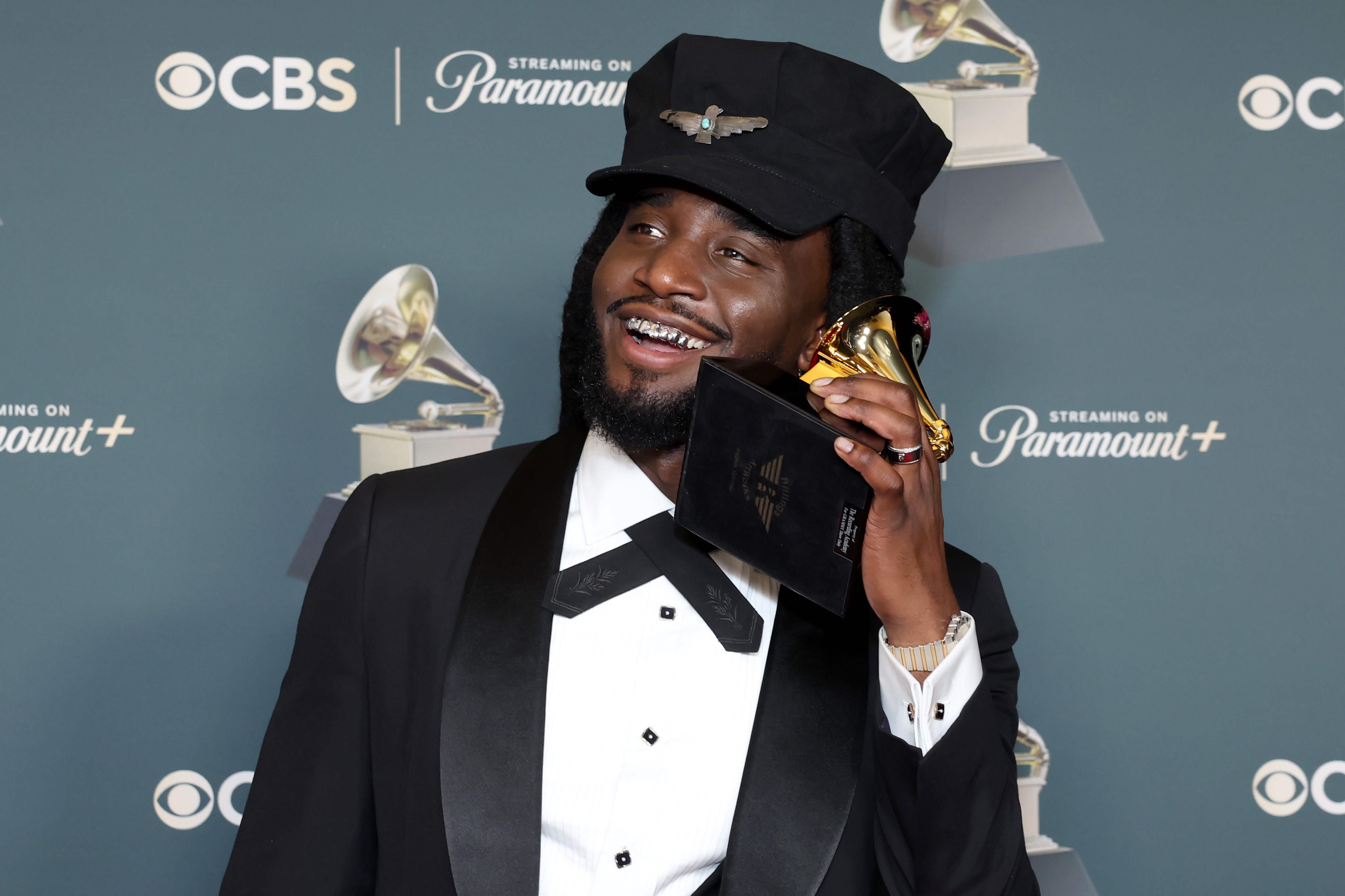 Person joyfully holding a Grammy award, dressed successful  a ceremonial  suit   with a unsocial   chapeau  connected  a Grammy-themed backdrop