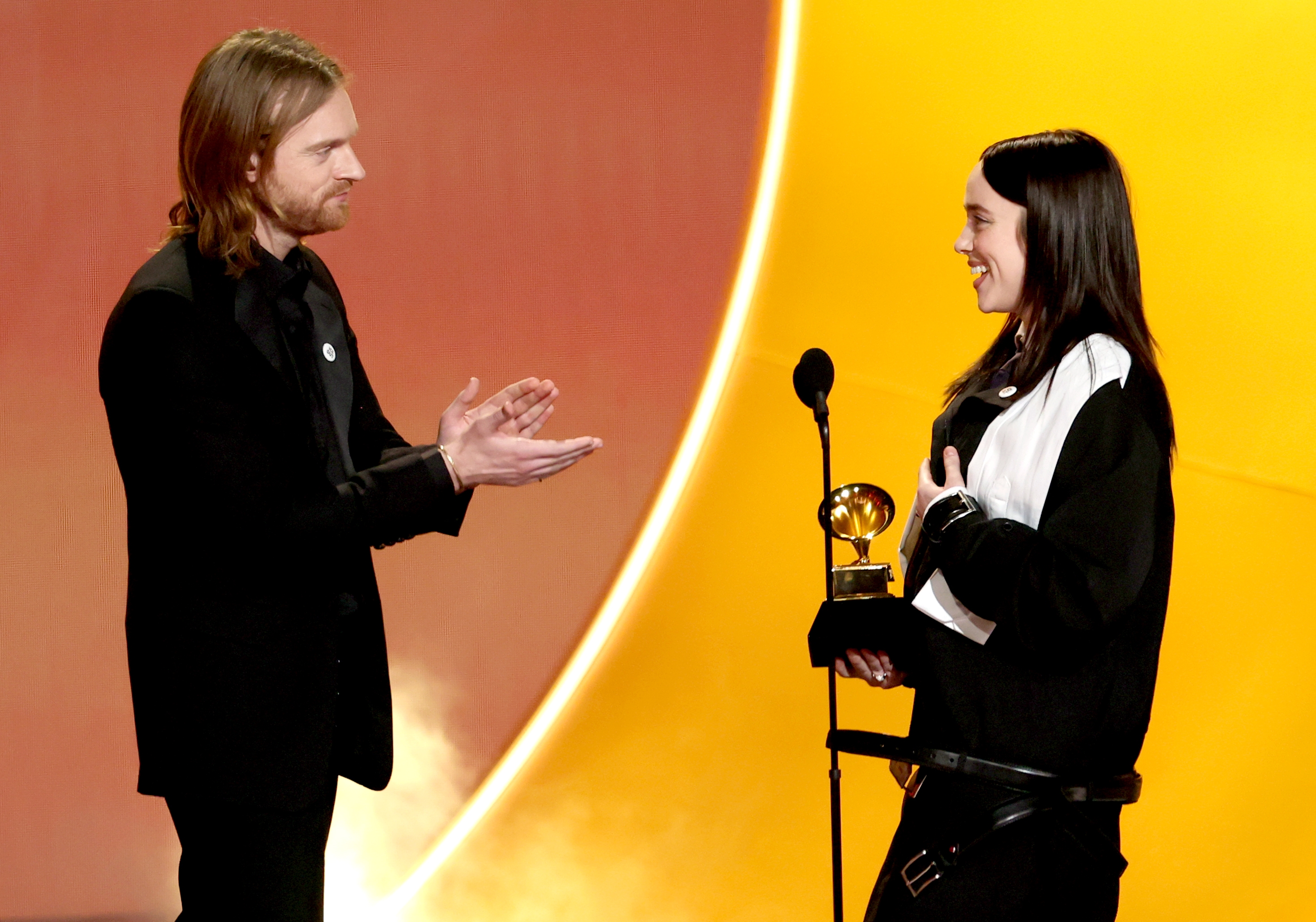 Two radical   connected  stage, 1  giving a Grammy grant  to the other. The grant  recipient wears a stylish, oversized outfit