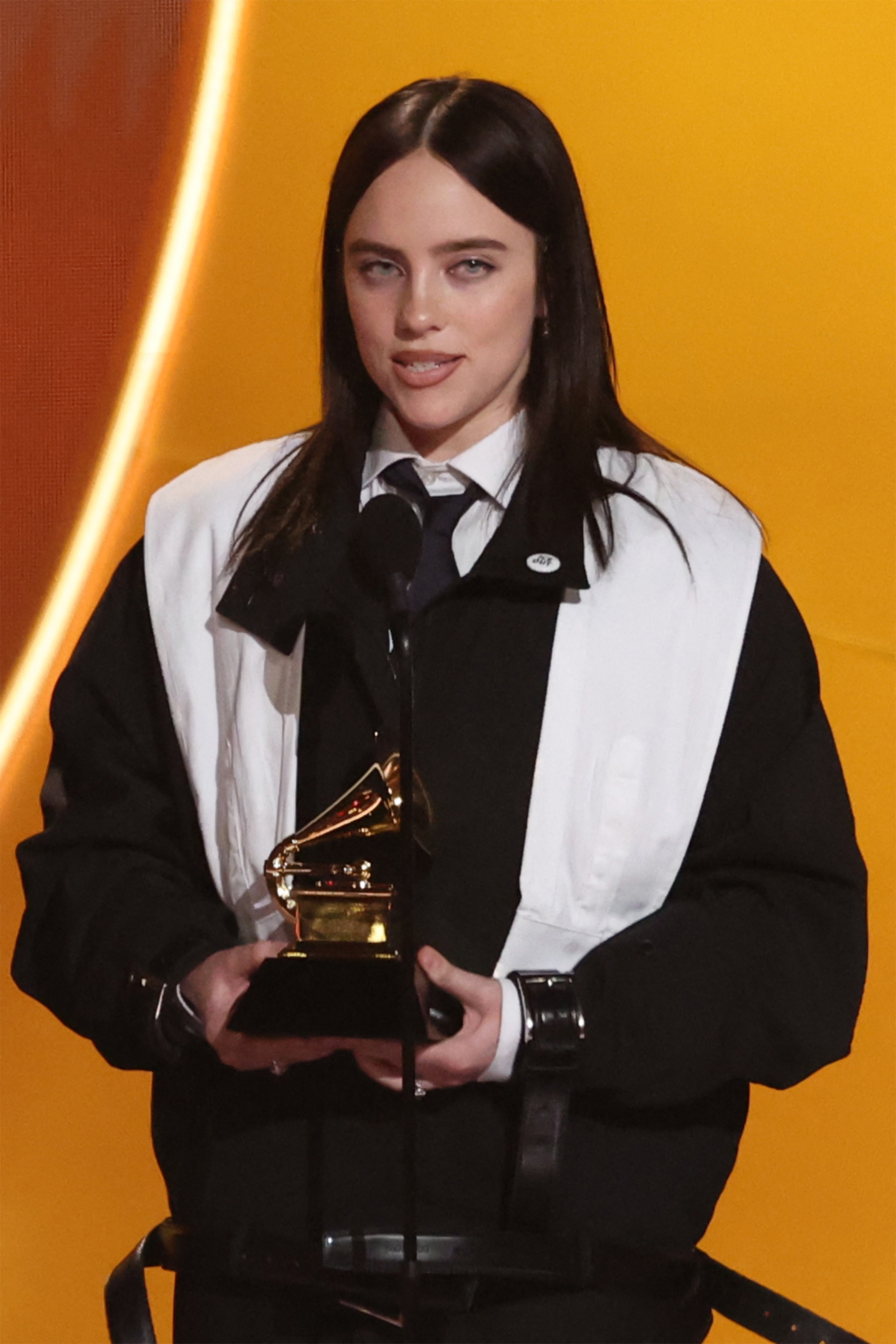 Person holding a Grammy trophy, dressed successful  a achromatic  and achromatic  overgarment   implicit    a achromatic  garment  with a tie, speaking astatine  a podium