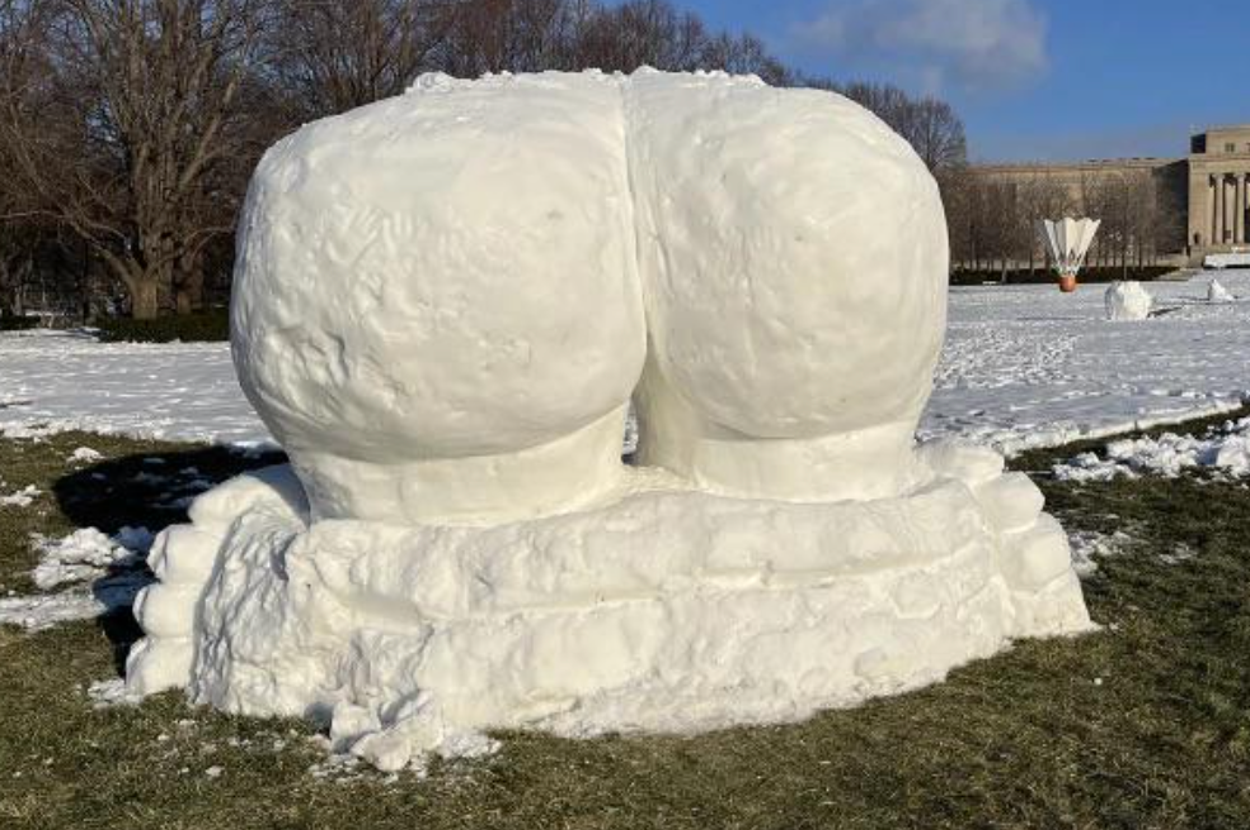 Snow sculpture resembling a large pair of human buttocks, situated on a grassy area with trees and a building in the background