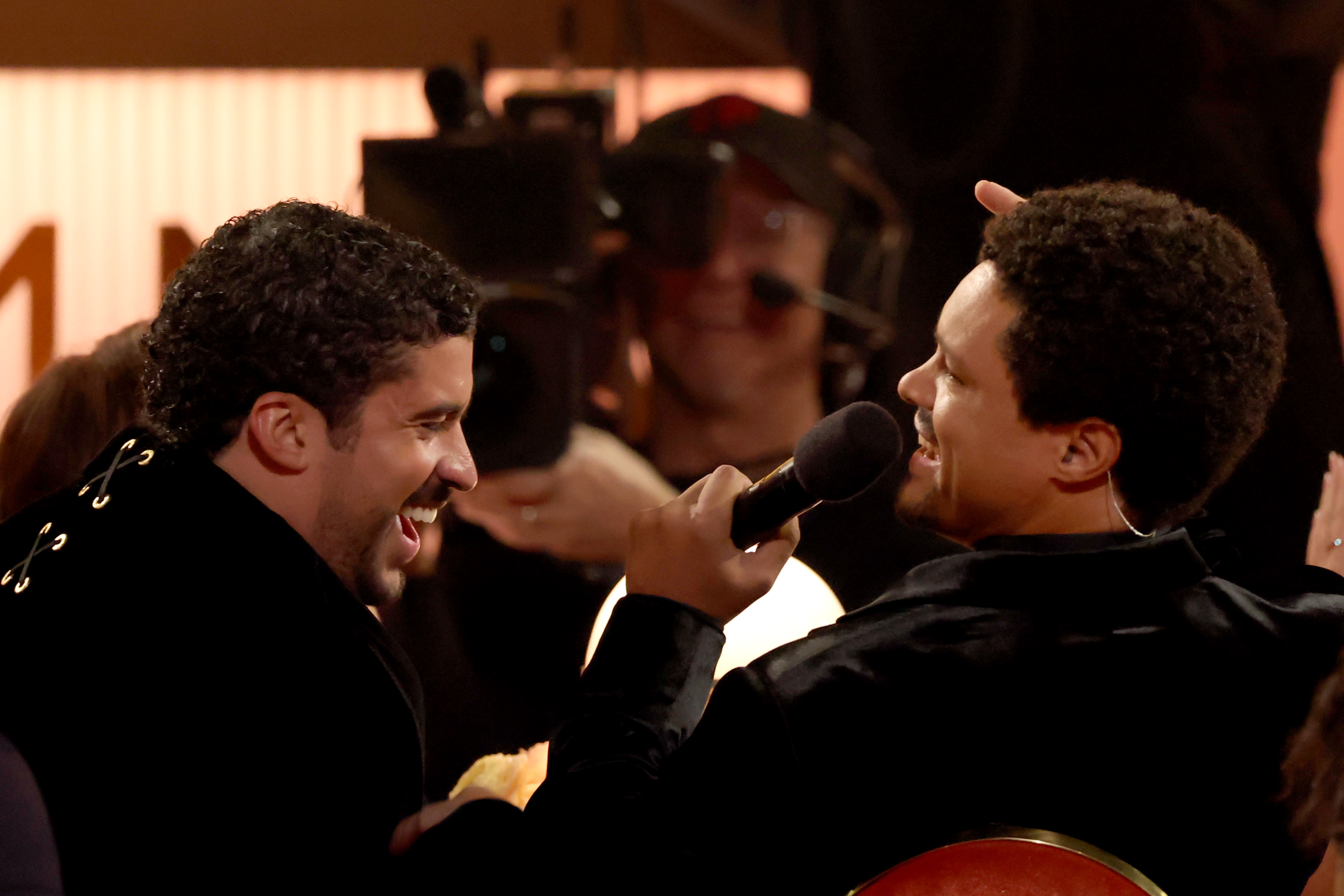 Two men successful  lively speech  astatine  an event, 1  holds a microphone. A camera idiosyncratic   is disposable   successful  the background