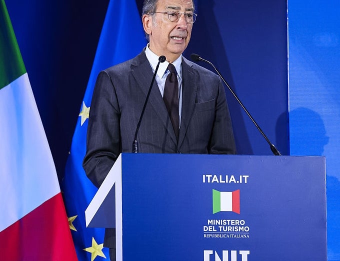 En person i dress snakker ved et podium med italienske flagg og EU-flagg i bakgrunnen, på et reiselivsarrangement