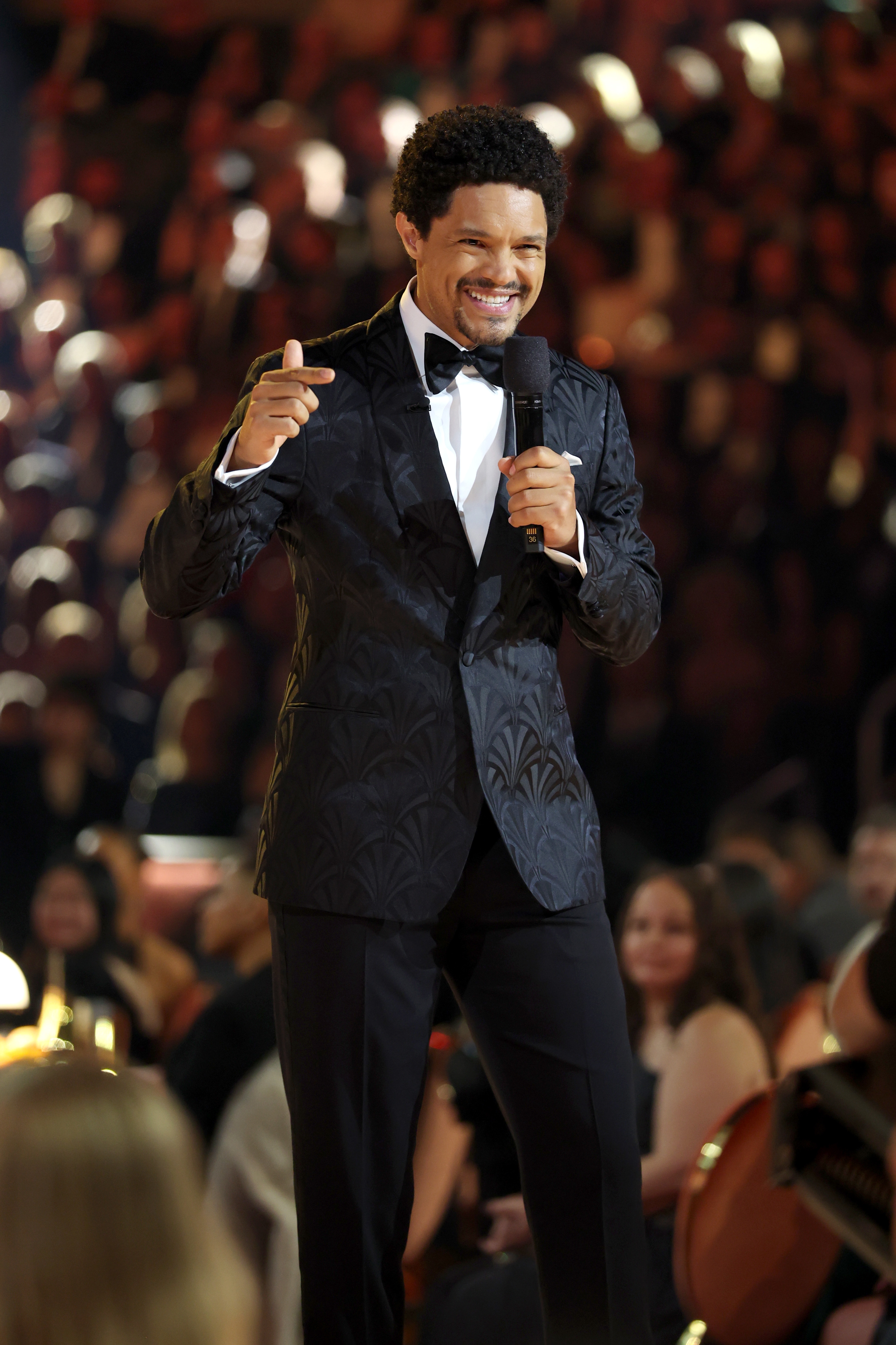 Mann i teksturert dress og sløyfe på scenen, snakker i en mikrofon. Publikum i bakgrunnen