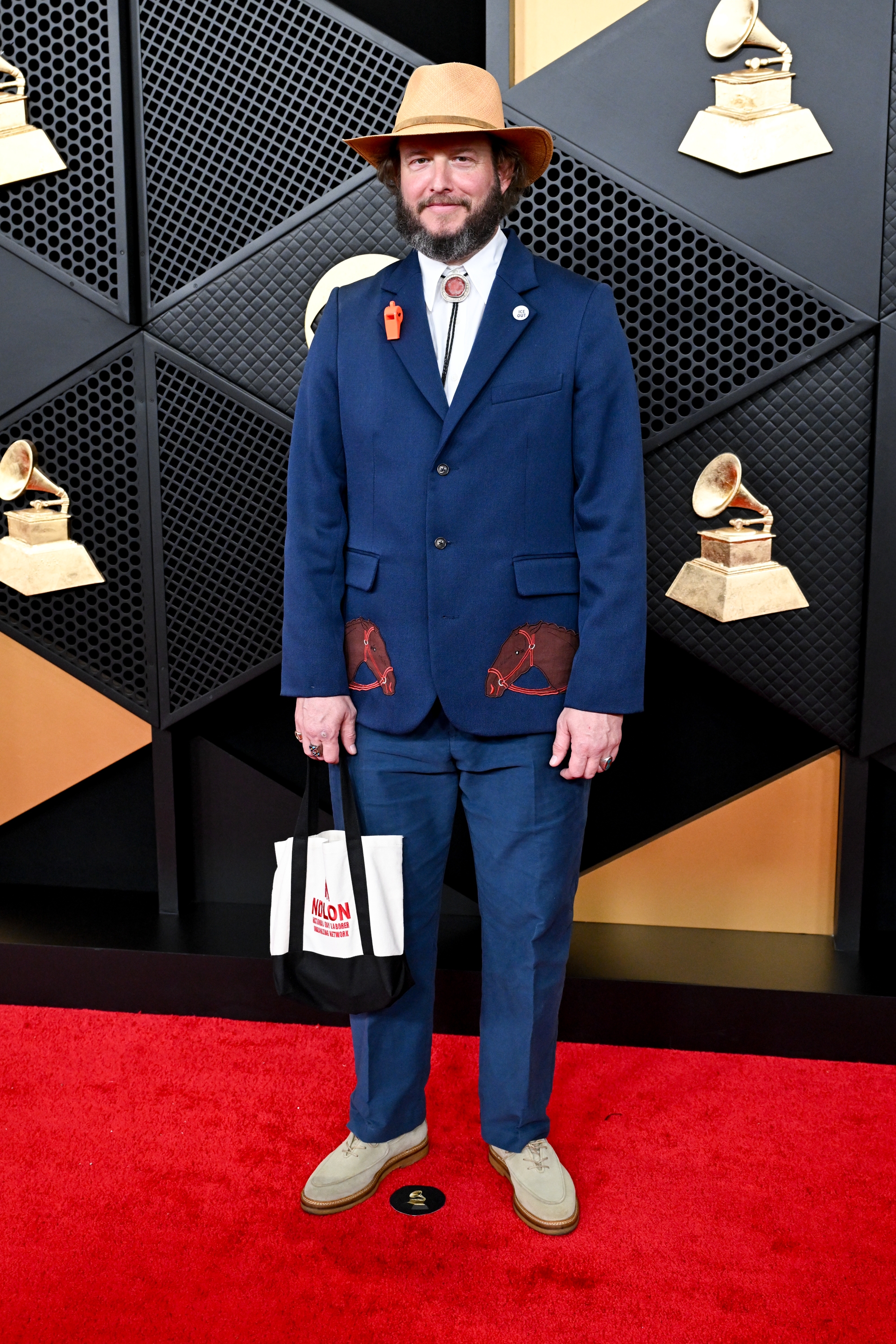 Man successful  a bluish  suit   with unsocial   spot  designs, wearing a chapeau  and holding a tote bag, stands connected  a reddish  carpet with Grammy Awards backdrop