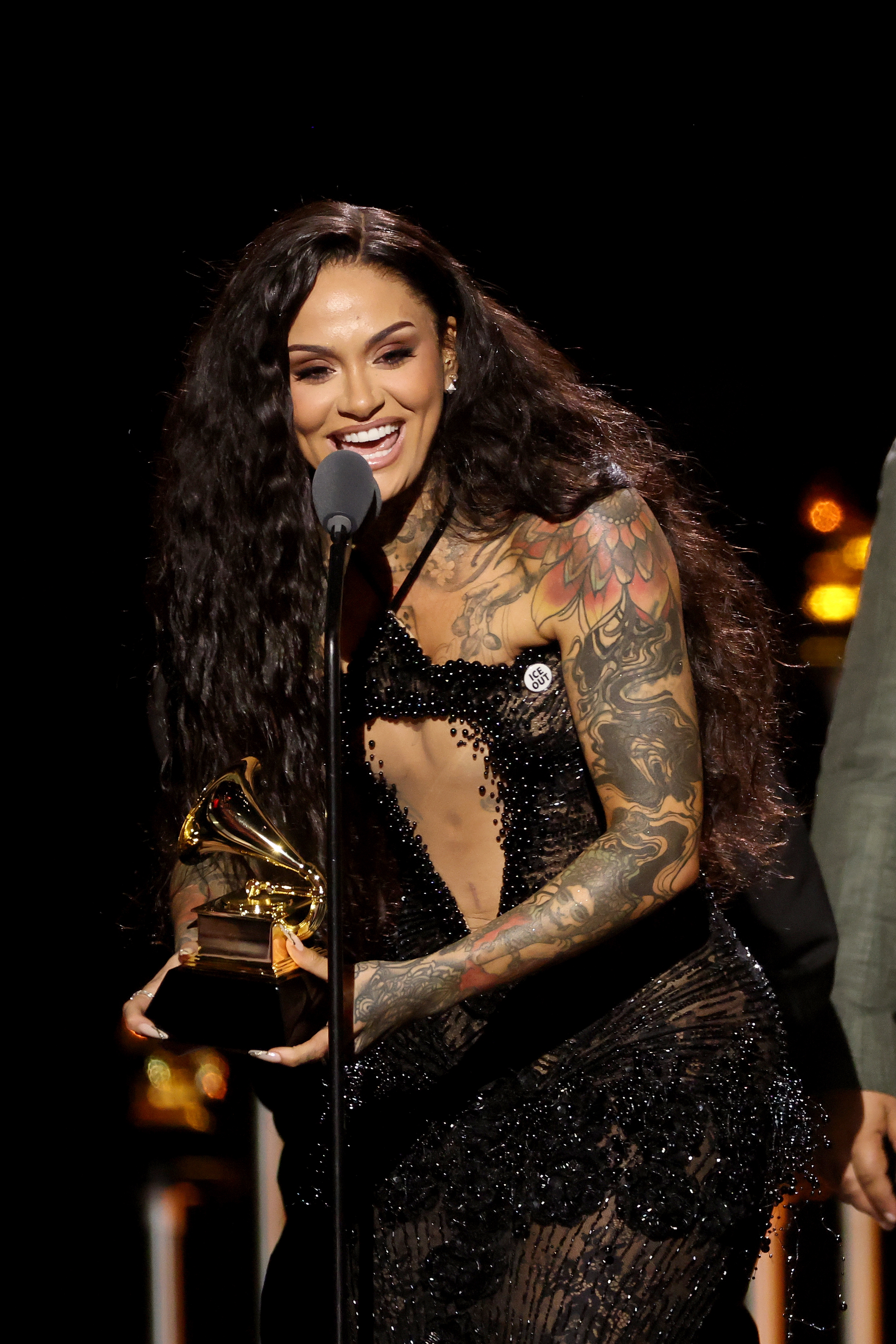 Person with agelong  hairsbreadth  and tattoos wearing a sparkly, embellished dress, holding a Grammy grant  and speaking connected  stage