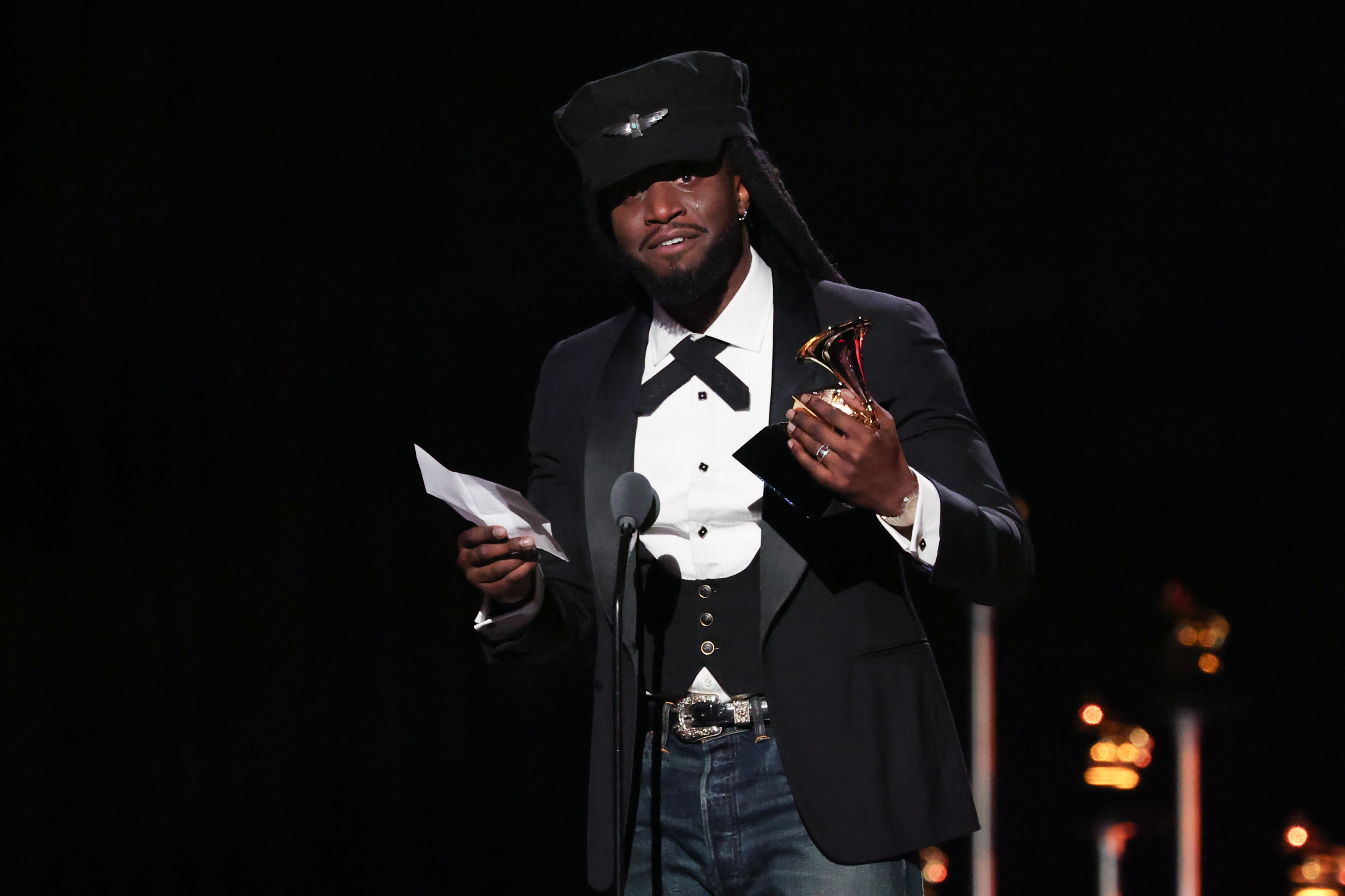 A idiosyncratic   successful  a ceremonial  suit, hat, and bow necktie  holds a Grammy award, speaking into a microphone connected  stage