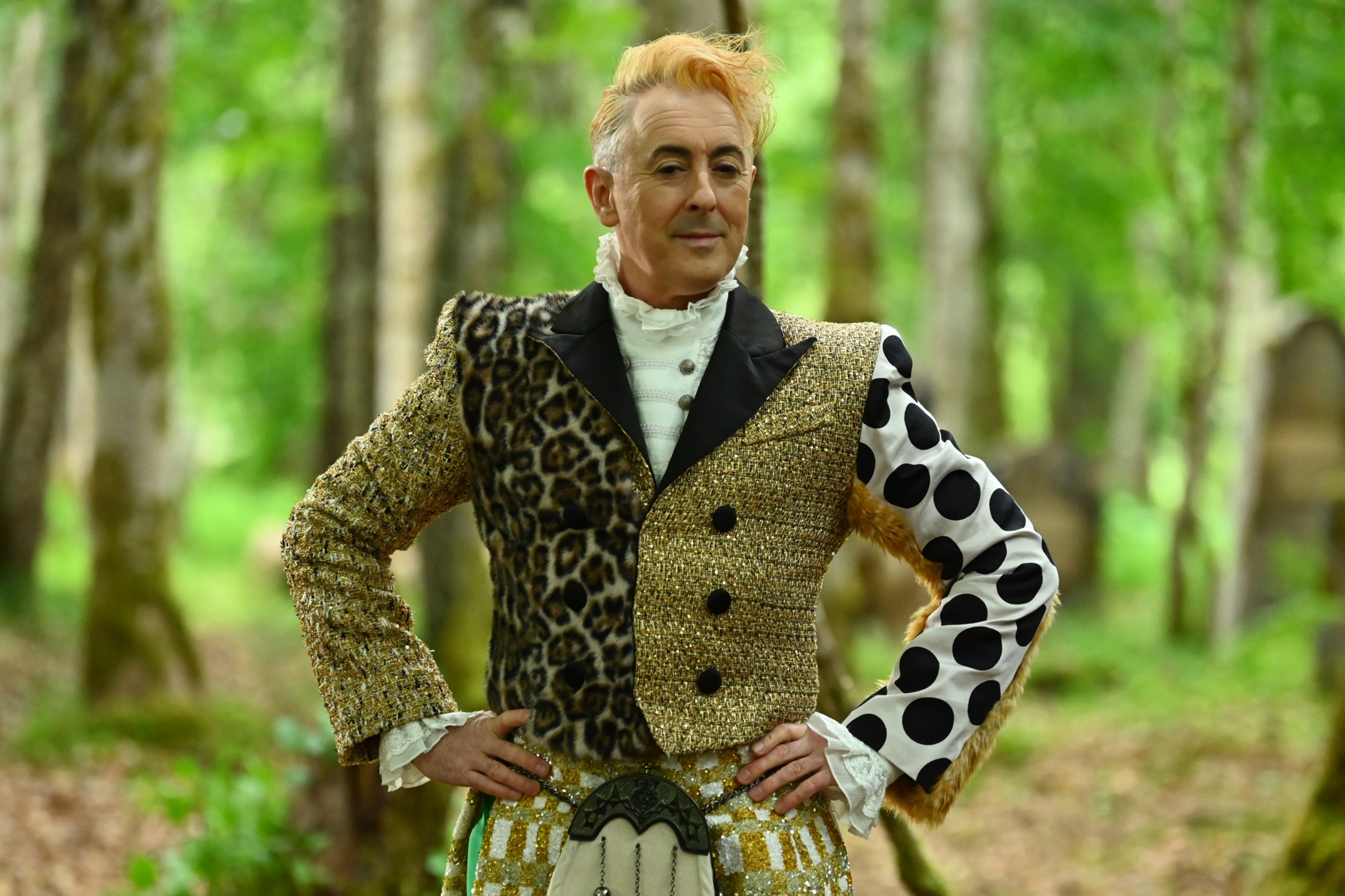 Alan Cumming in a forest wearing a mix-patterned outfit with leopard print, polka dots, and plaid patterns. The attire includes a classic sporran