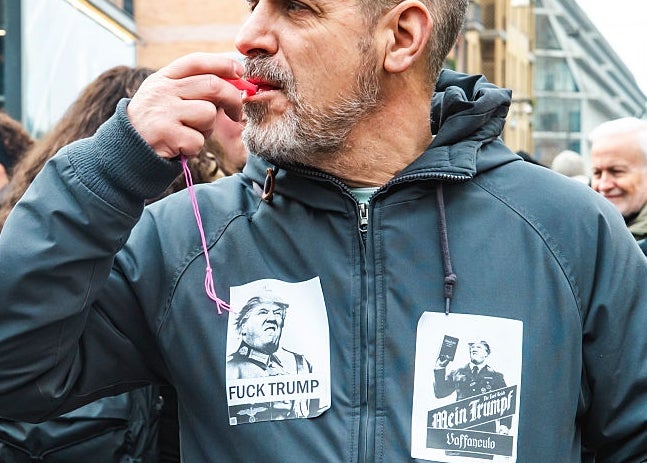 Person i en folkemengde som har på seg en grå jakke med anti-Trump-klistremerker, blåser i fløyte under en gateprotest