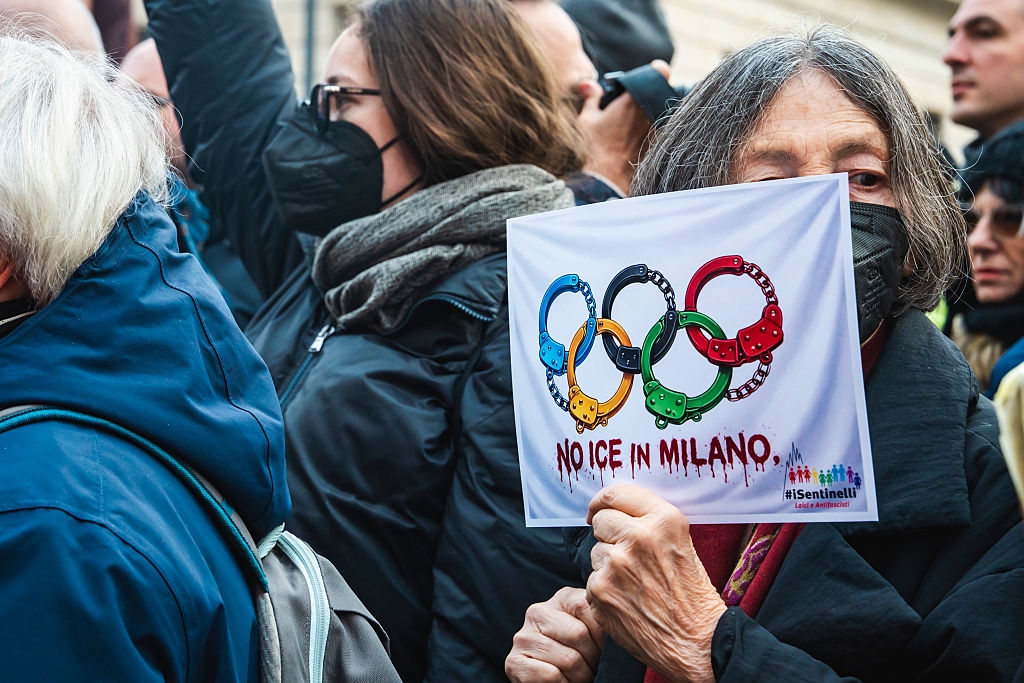 Person som holder et skilt med olympiske ringer formet som håndjern og leser «Ingen is i Milano»; under en protest. Andre står i nærheten, iført masker
